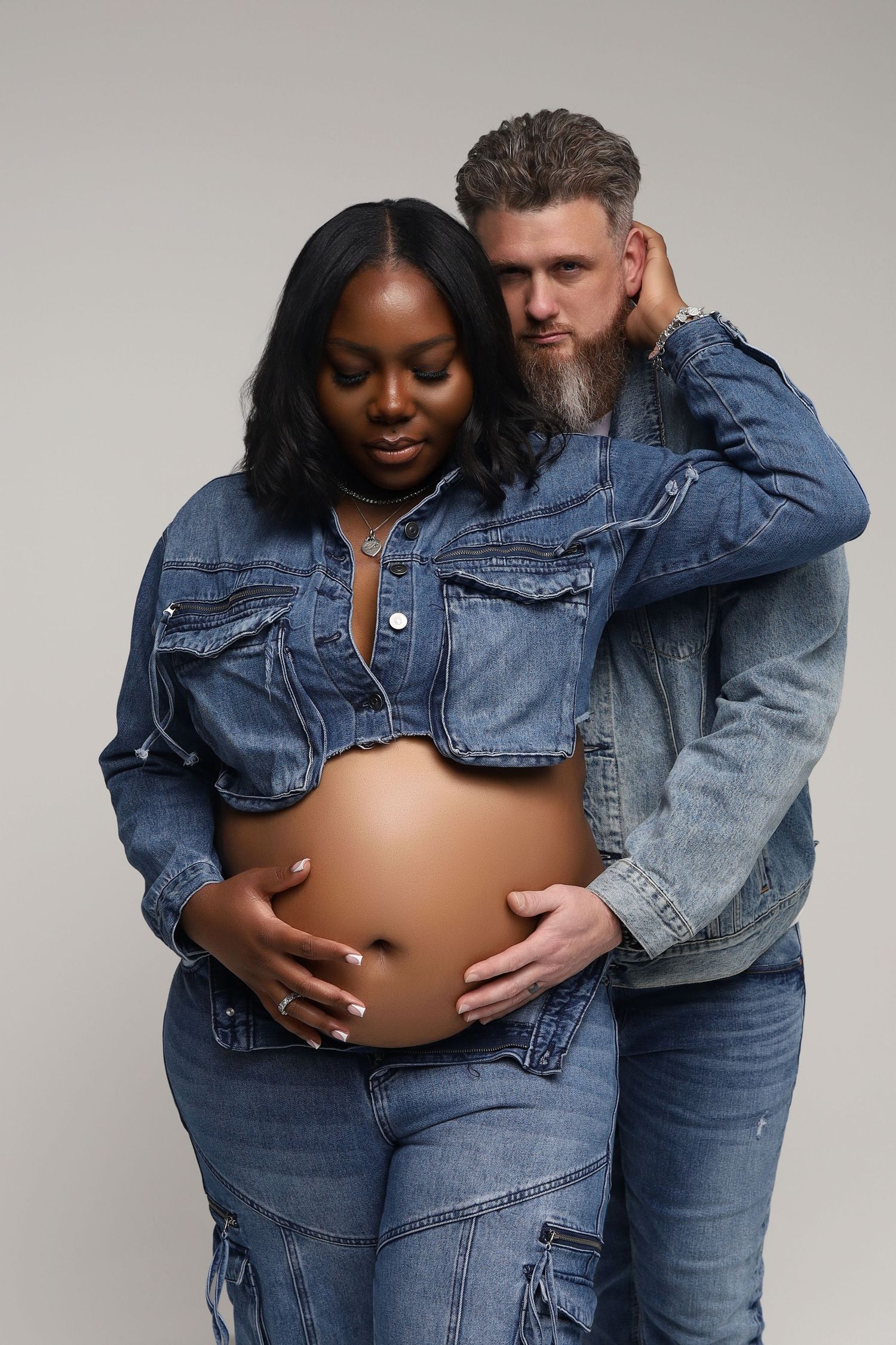 Interracial couple in denim outfits pose together, woman showing pregnant belly in studio.