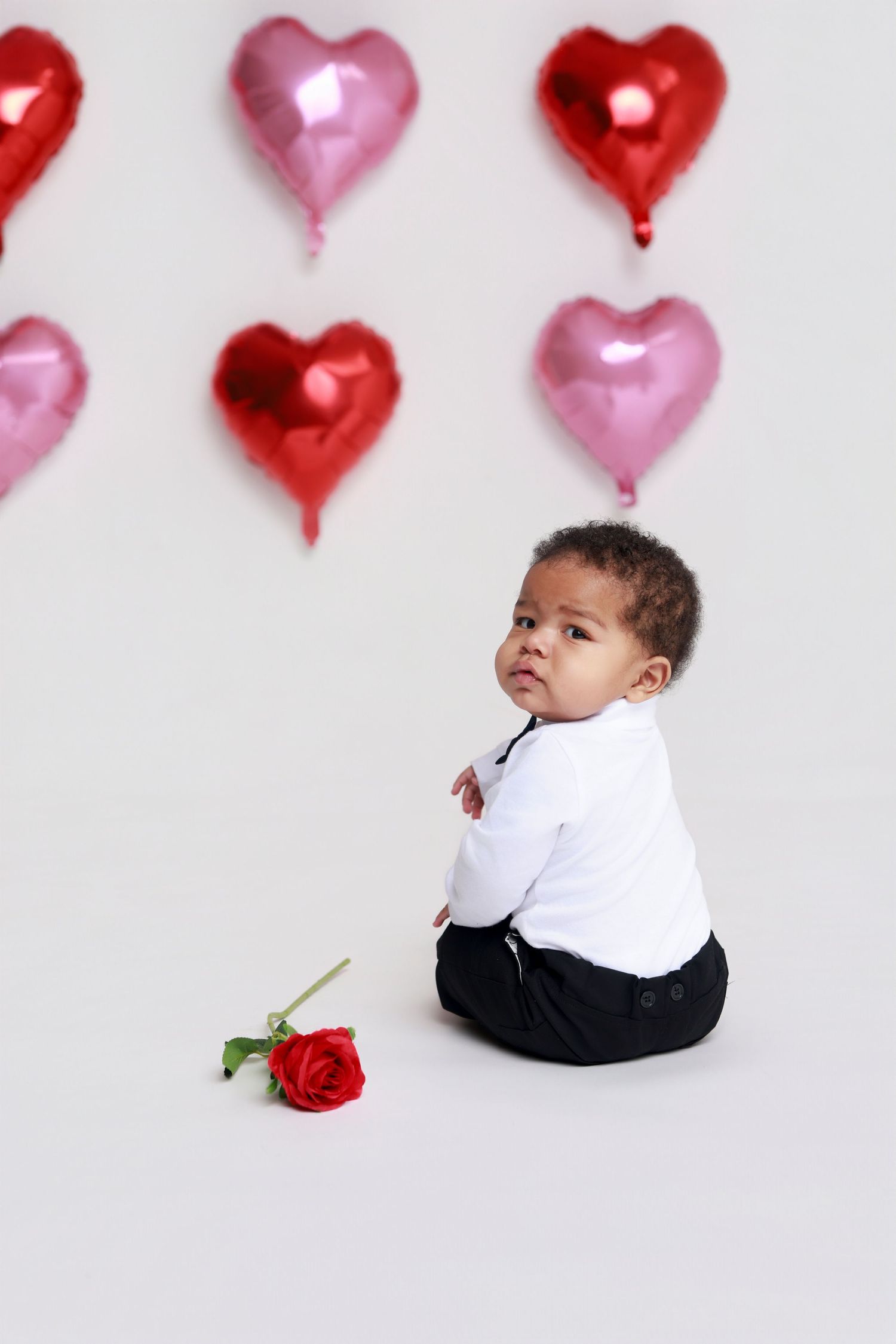 An infant in a white shirt and black pants sits near a red rose with heart balloons behind them.