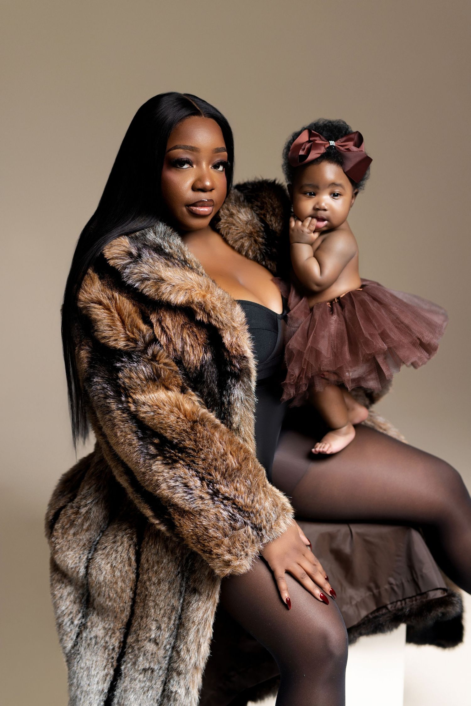 A Black woman in a fur coat holds an infant in a brown tutu in a professional studio portrait.