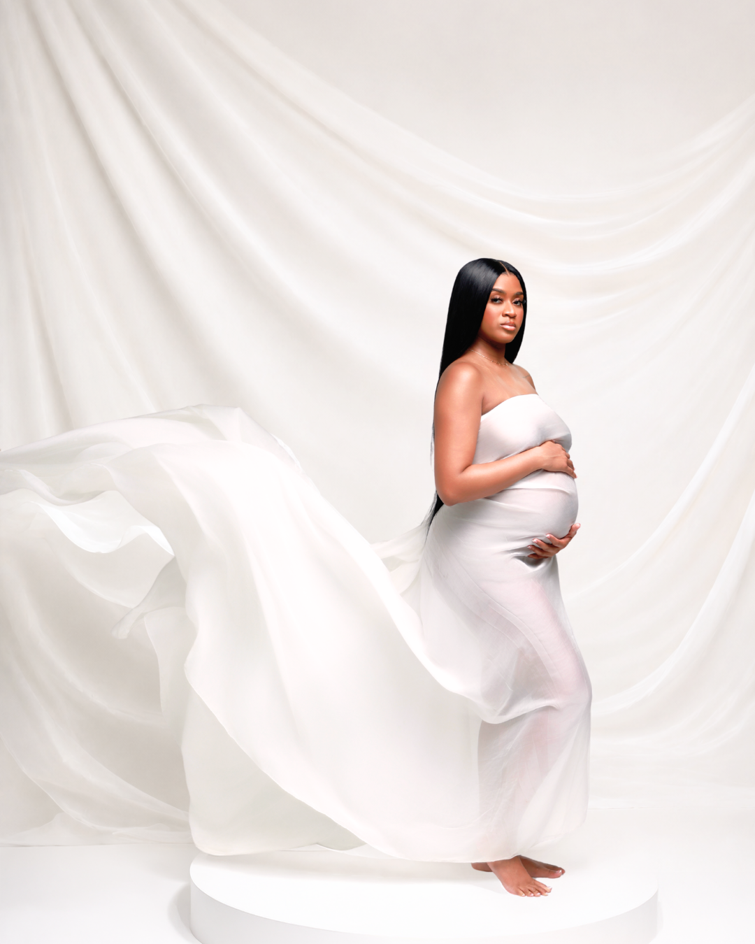 Elegant pregnant woman in flowing white fabric dress poses gracefully on white pedestal in studio.