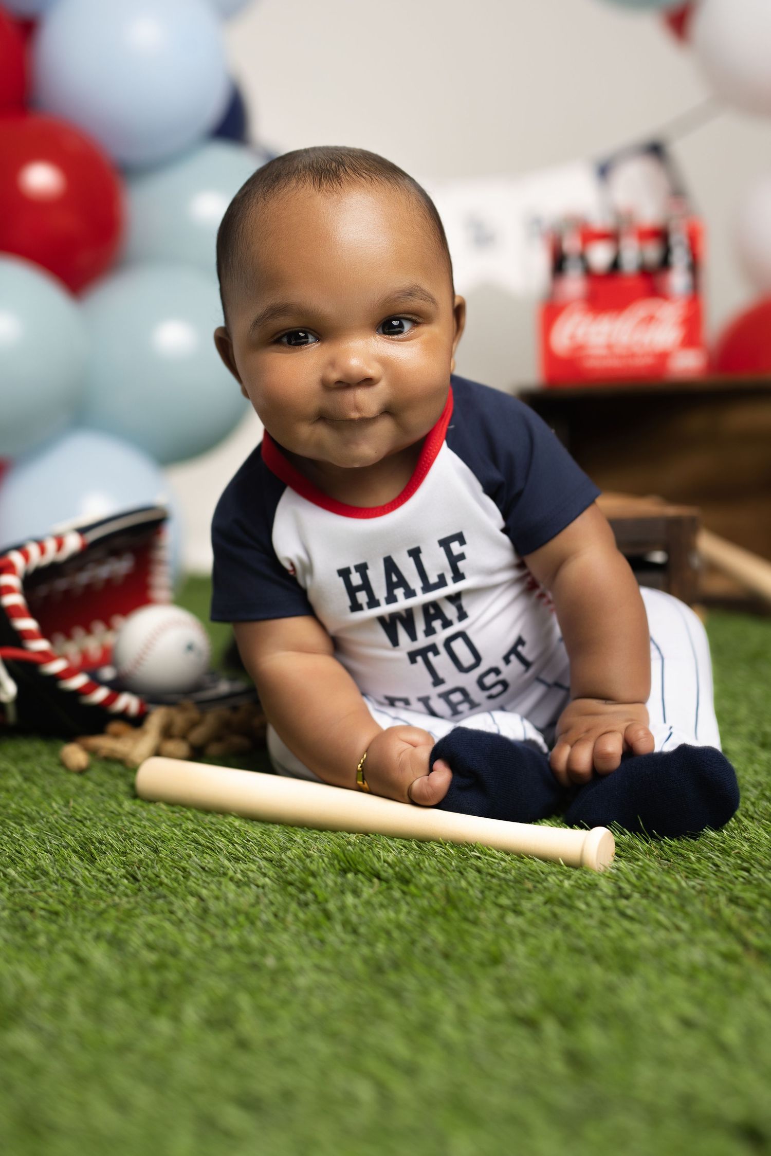 Smiling baby boy in baseball outfit sits on turf with bat and glove for sports photo.