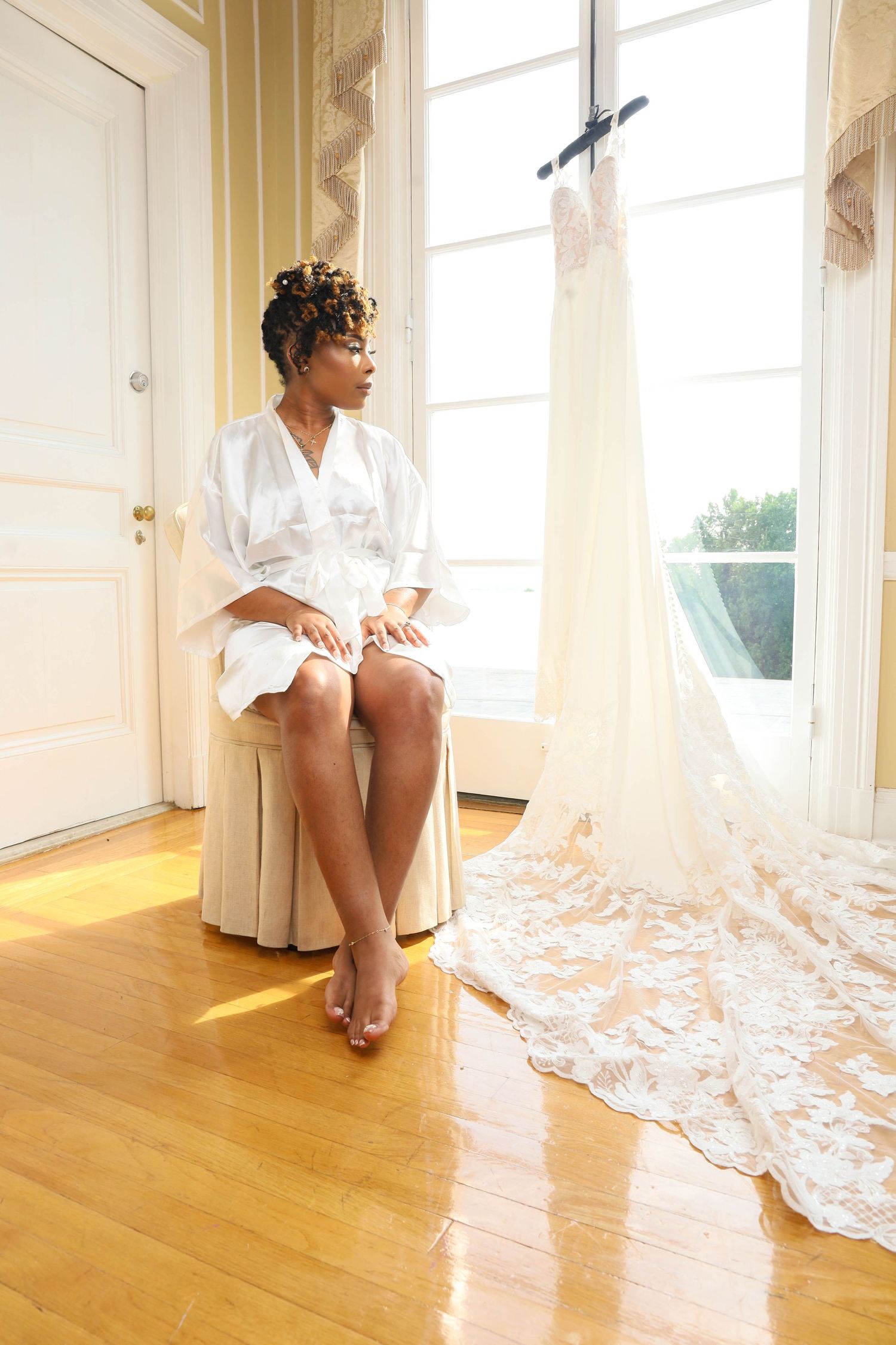 Bride in white robe sits beside her lace wedding dress hanging by a sunlit window.