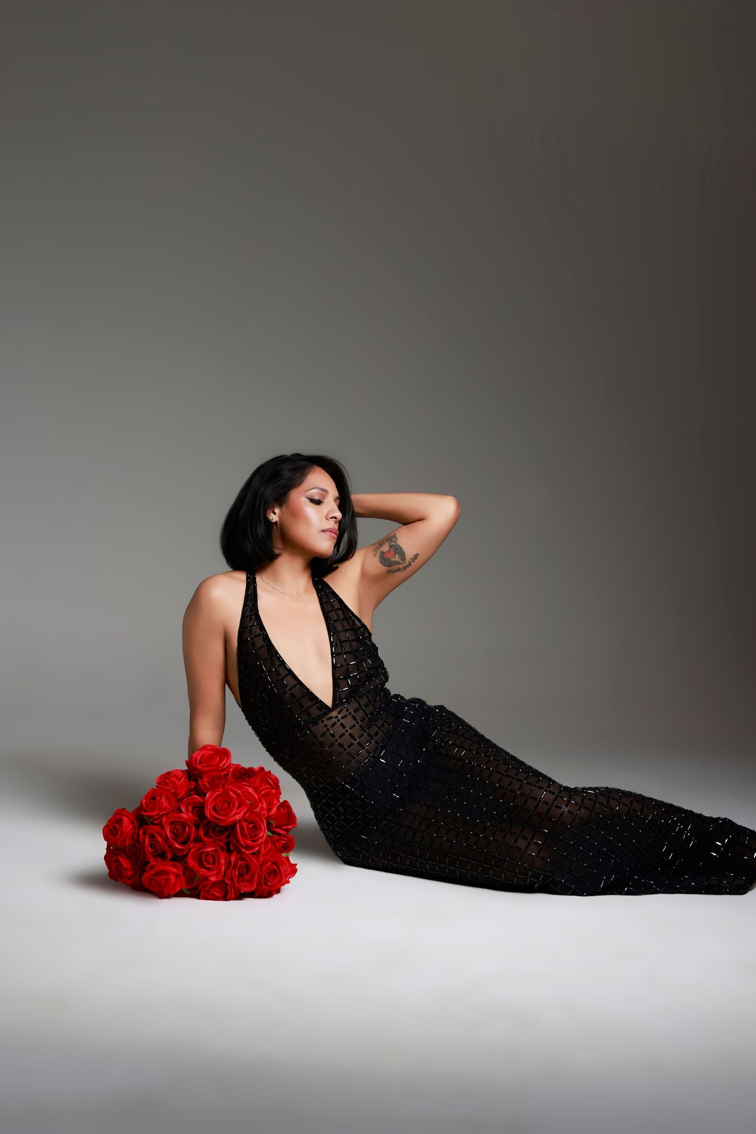 A woman in a black sequined gown reclines on the floor beside a bouquet of red roses.