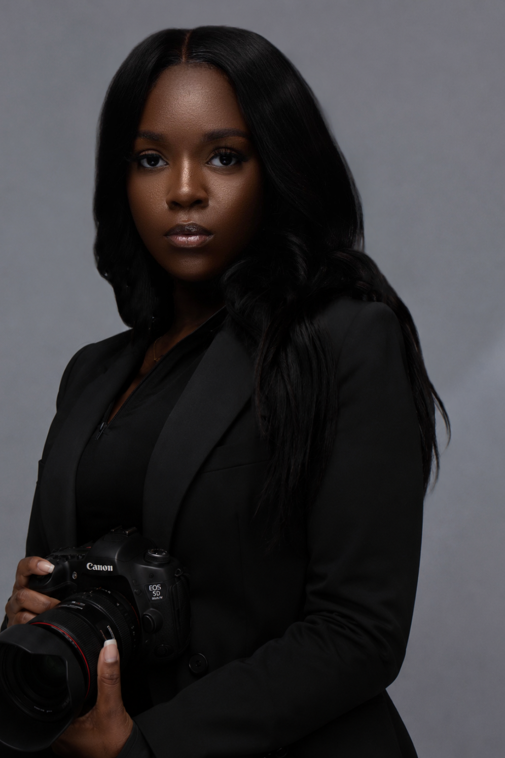 Young Black woman in black blazer holding Canon camera against gray background.