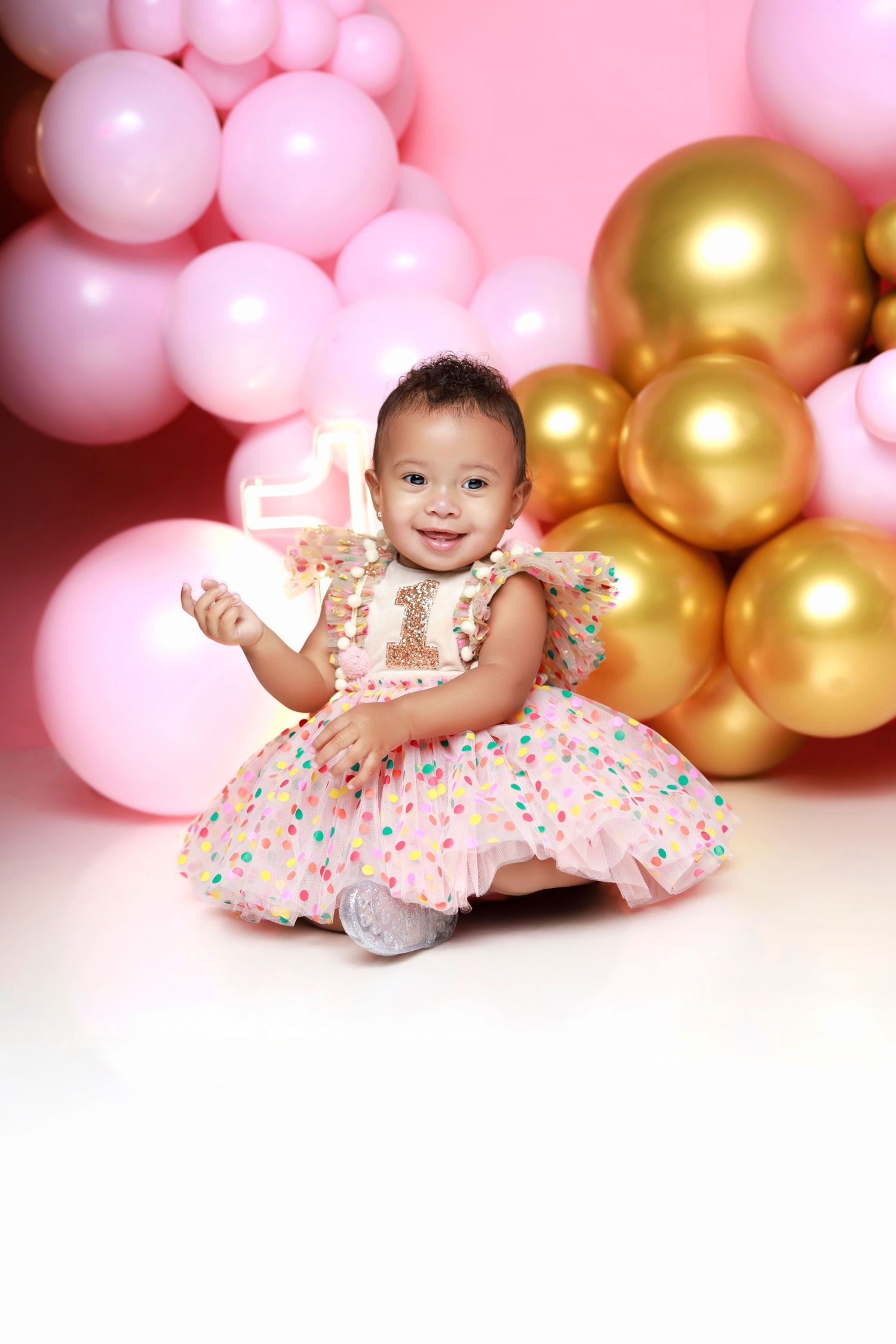 Happy baby girl in polka dot first birthday dress poses with pink and gold balloons.