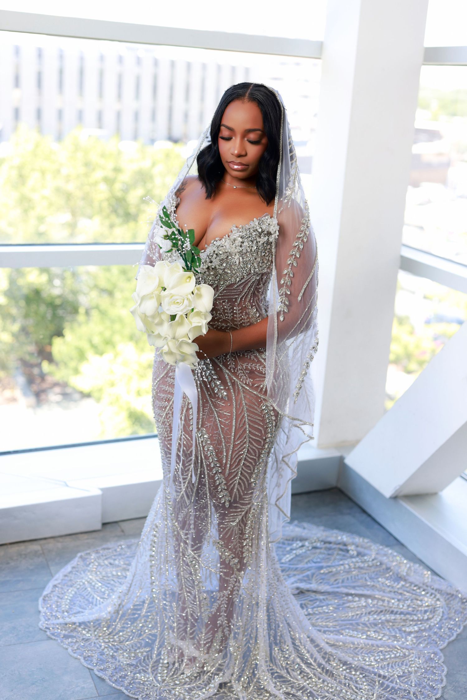 Bride in stunning beaded sheer gown holds white bouquet while wearing long veil by bright window.