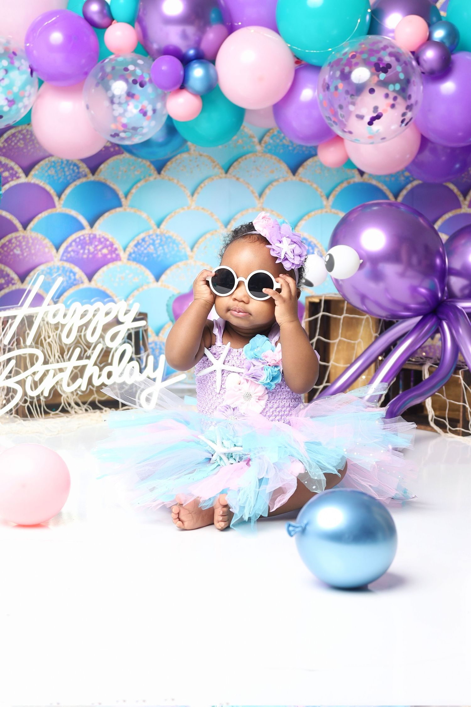 Baby girl in mermaid tutu dress with sunglasses celebrates first birthday with purple balloons.