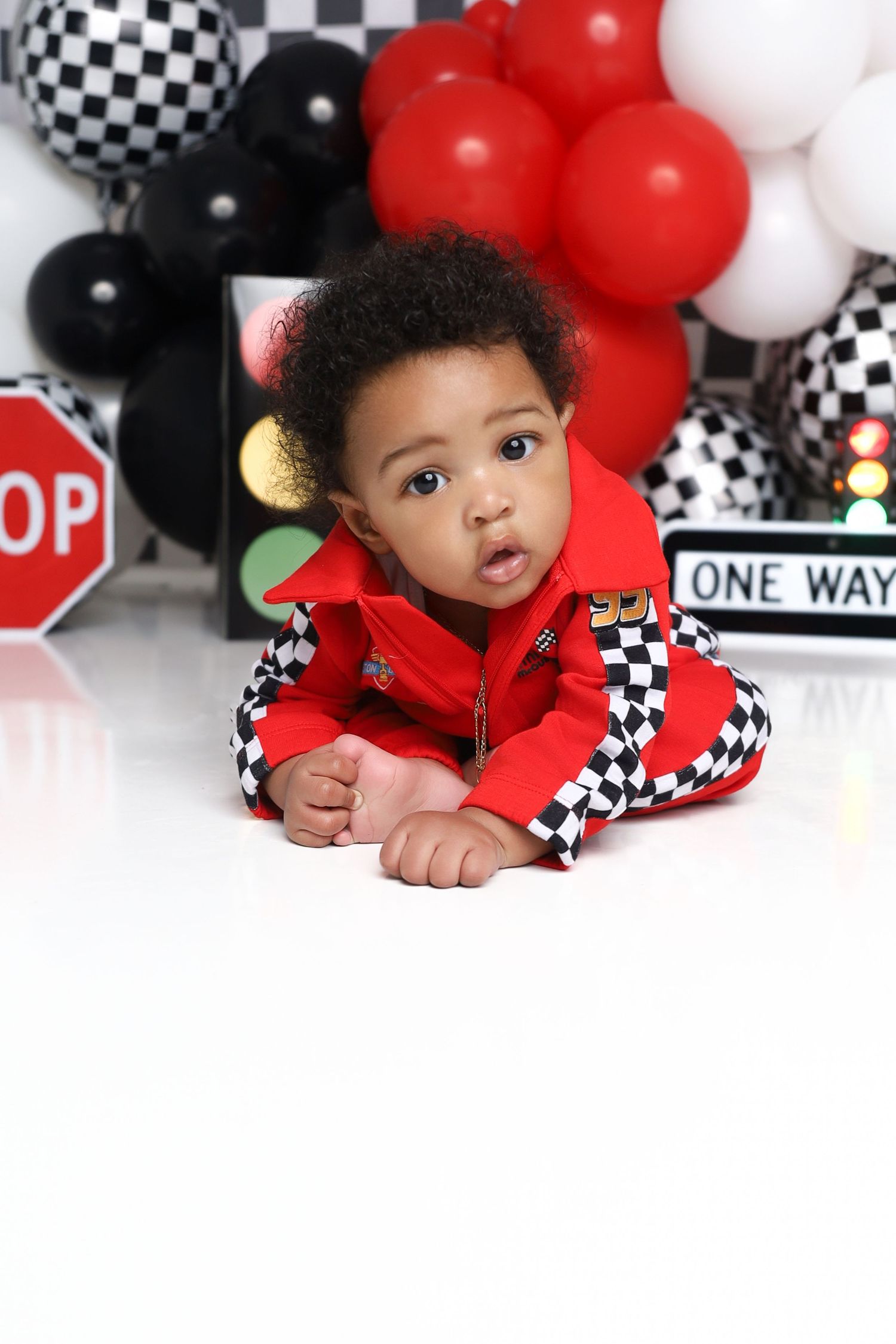A baby boy in a red checkered racing jacket lies on the floor with race-themed balloon decorations.