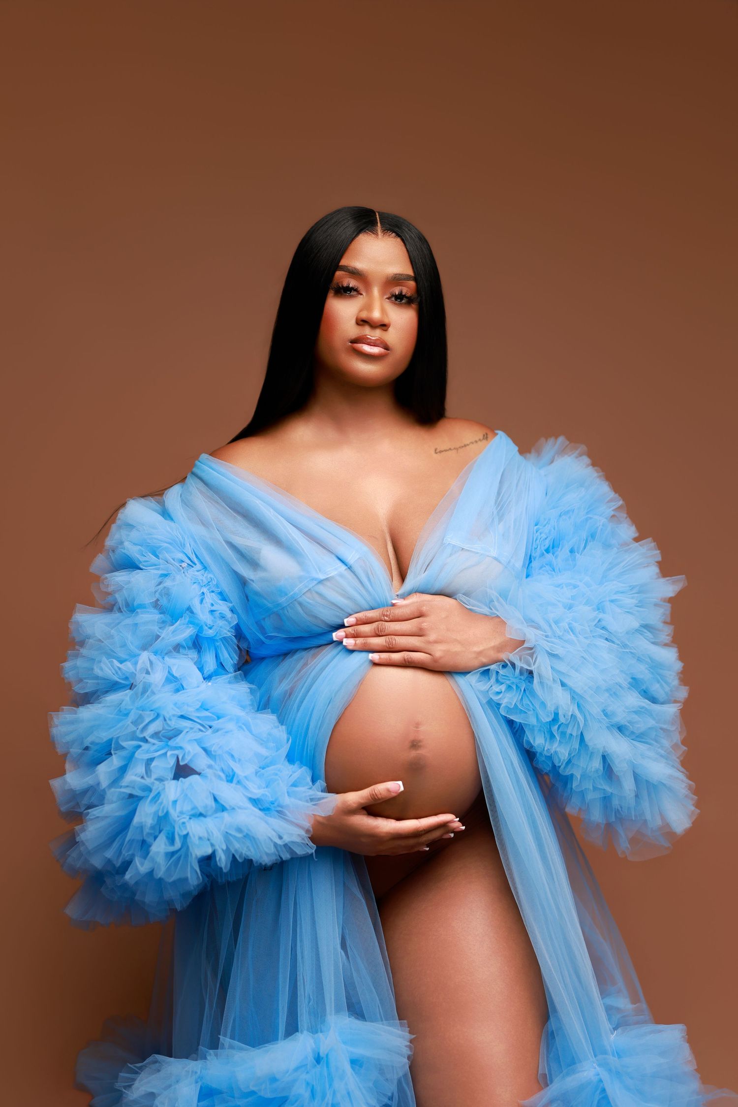 Pregnant woman in flowing blue ruffled tulle robe cradles baby bump against brown backdrop.