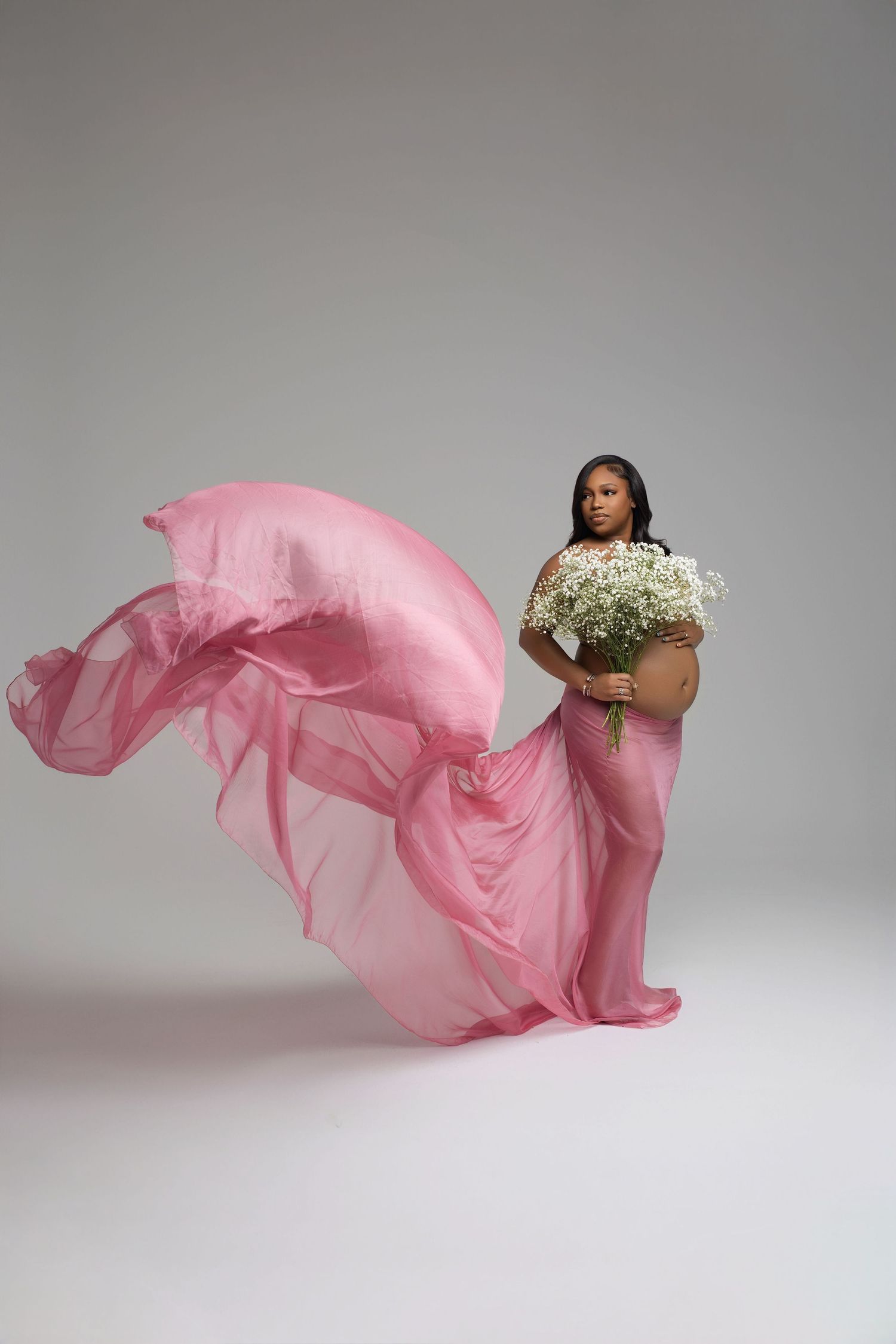 Pregnant woman holds white flowers while pink fabric flows dramatically in a studio maternity shoot.