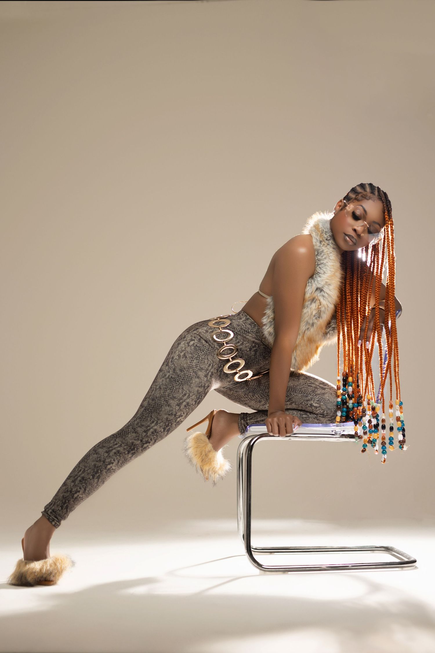 A model with long orange braids poses on a chrome chair wearing gray snakeskin leggings and fur vest.
