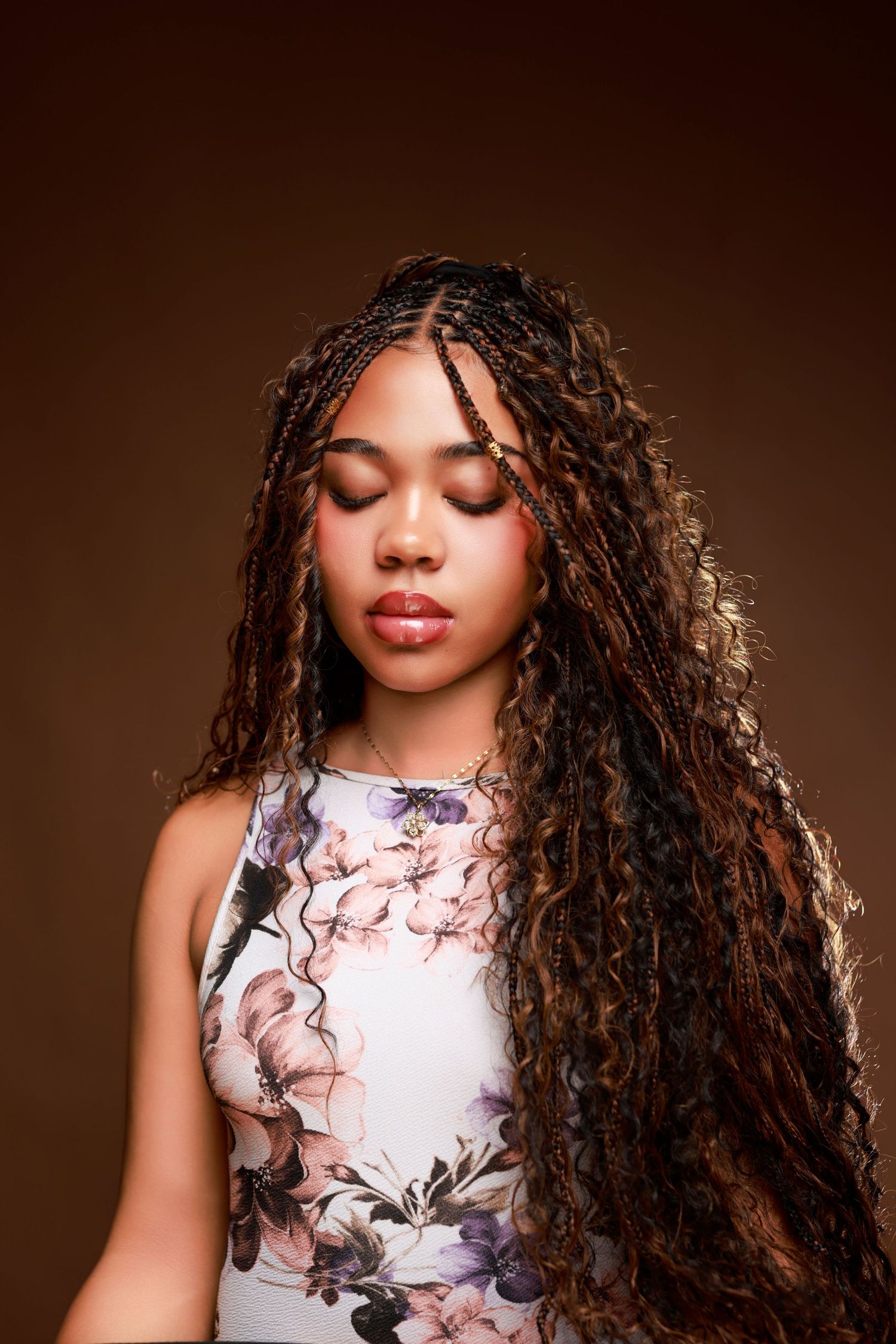 Young Black woman with long braided curly hair poses in floral top against a brown background.