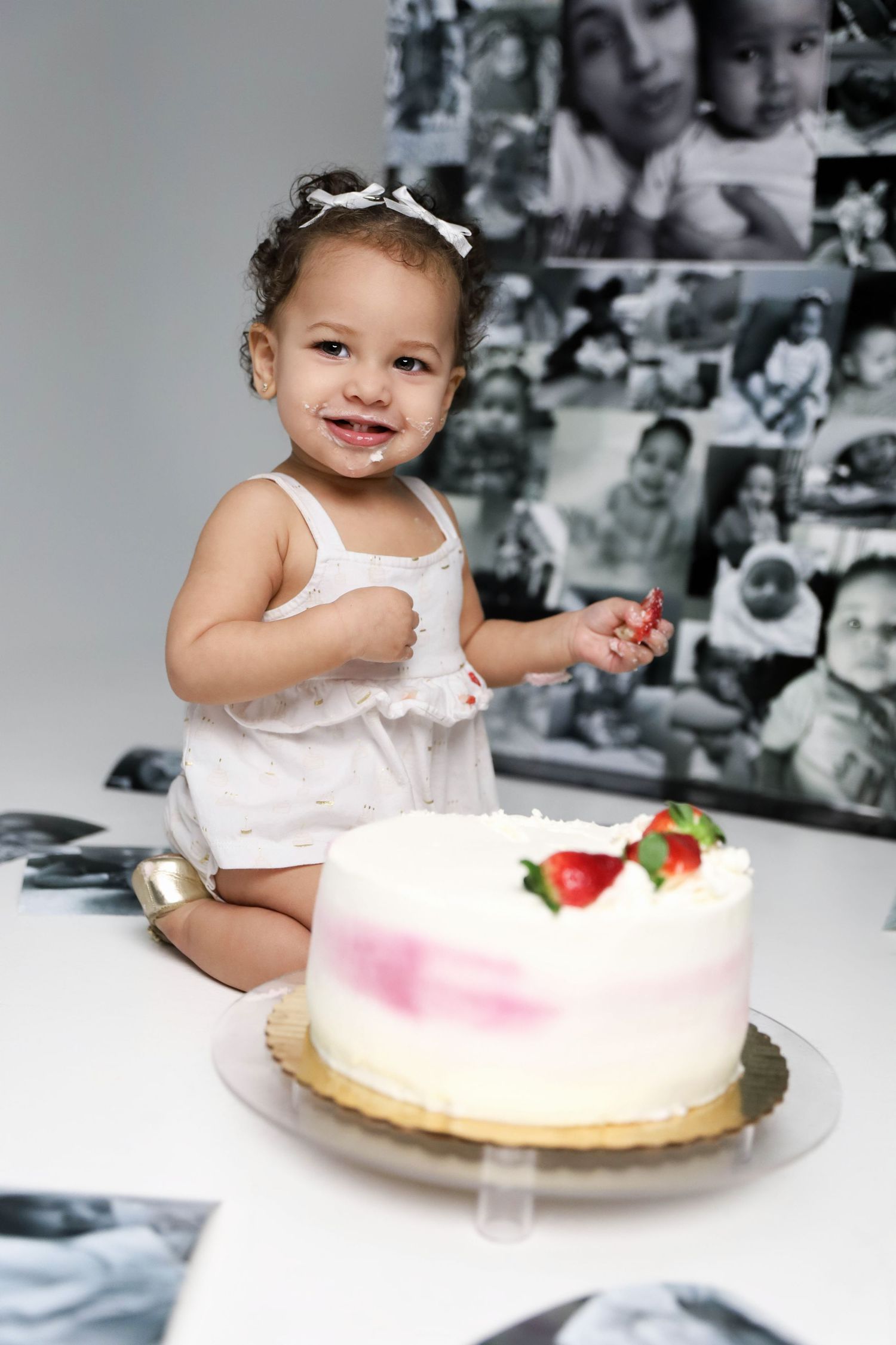 A smiling mixed-race toddler in white dress celebrates her first birthday with a strawberry cake.