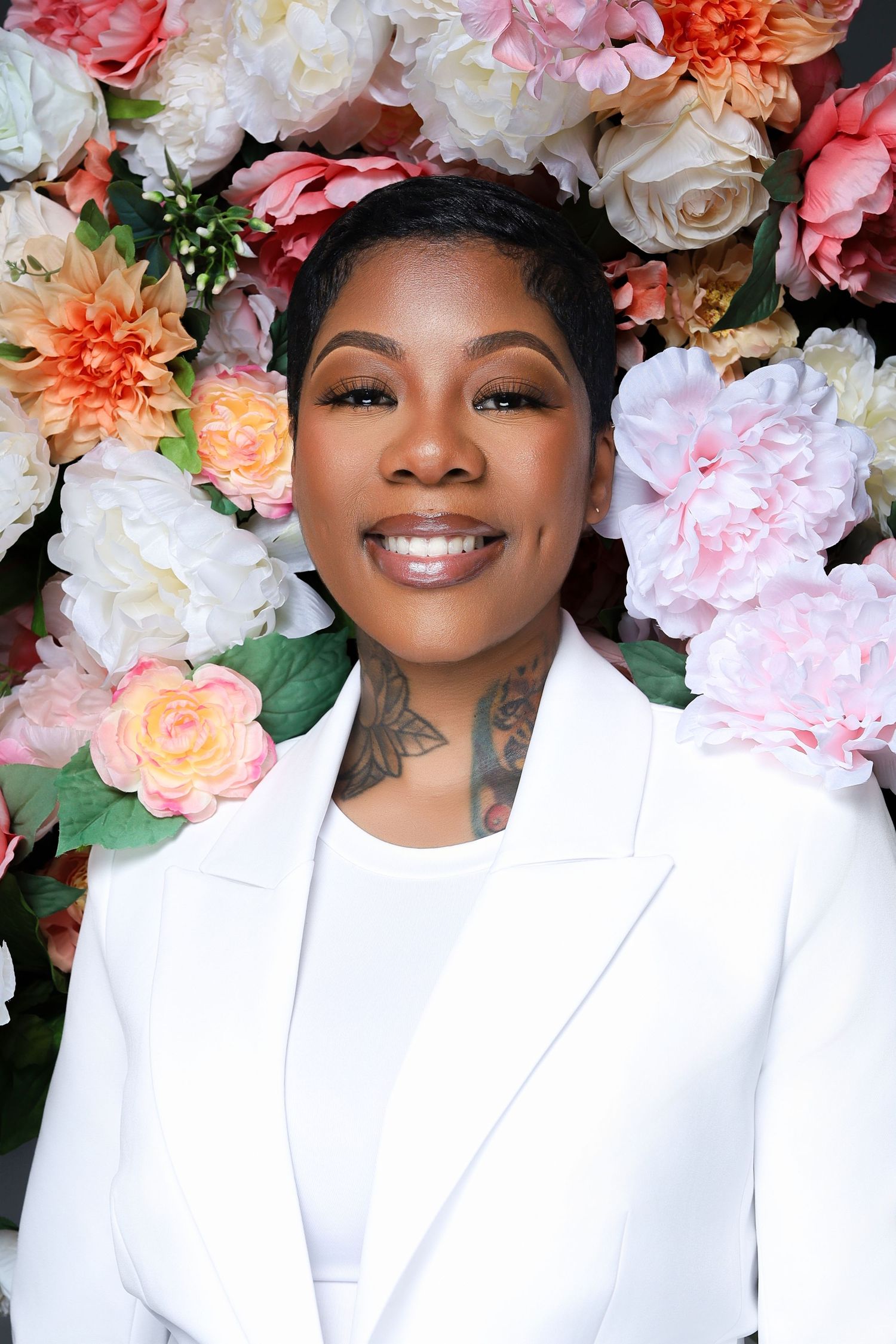 Smiling Black woman in white blazer with neck tattoo poses before a colorful floral backdrop.