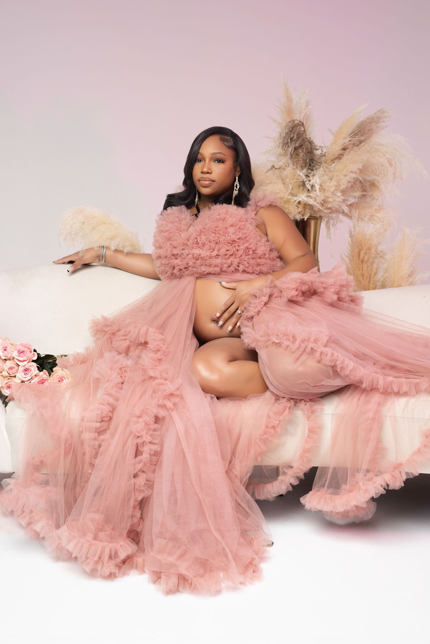Pregnant woman in pink ruffled tulle robe reclines on white sofa in elegant maternity portrait.