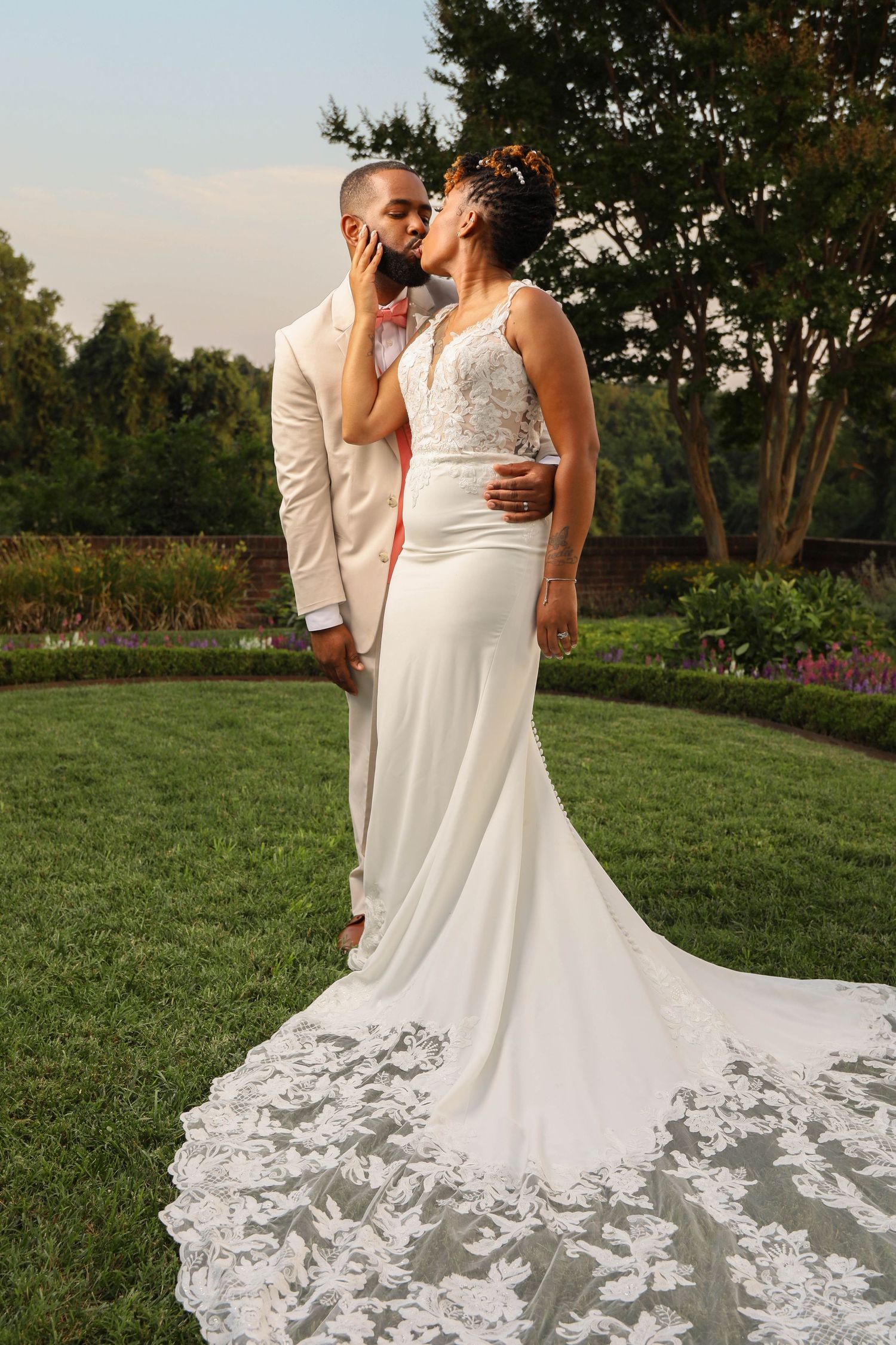 Bride and groom share a kiss in a lush garden, bride wearing a lace-train mermaid gown.