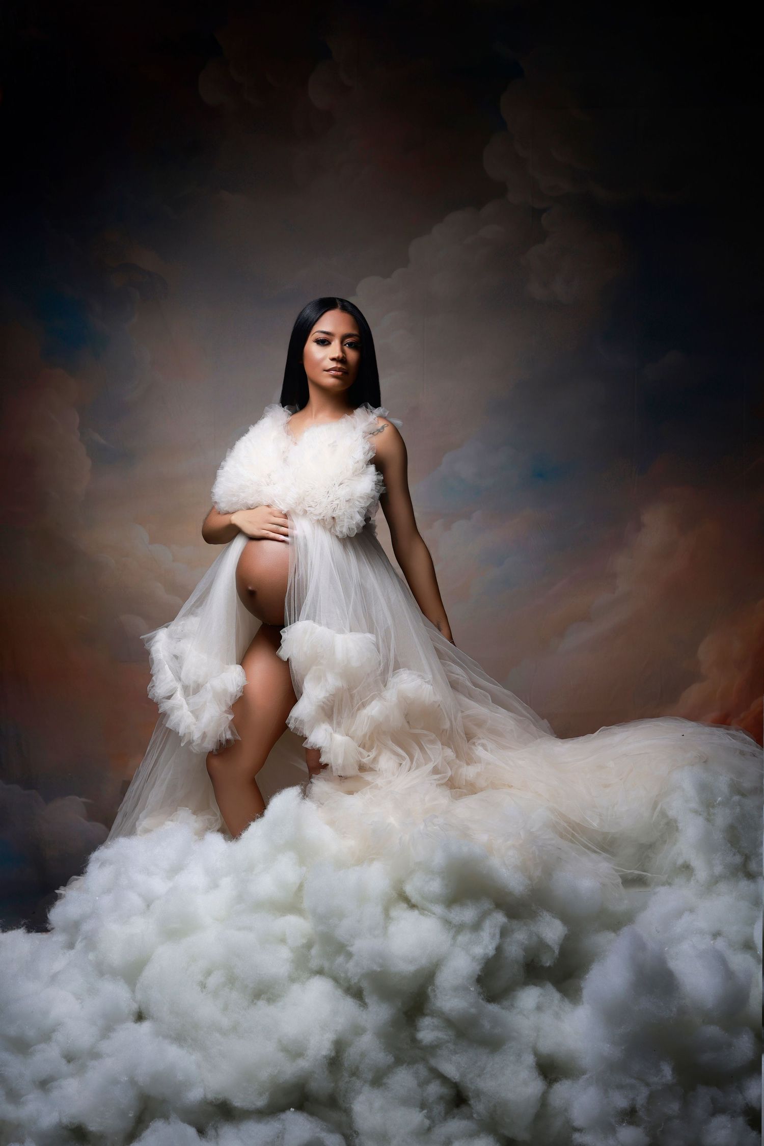 Pregnant woman in sheer white ruffled gown poses majestically on clouds with dramatic sky backdrop.