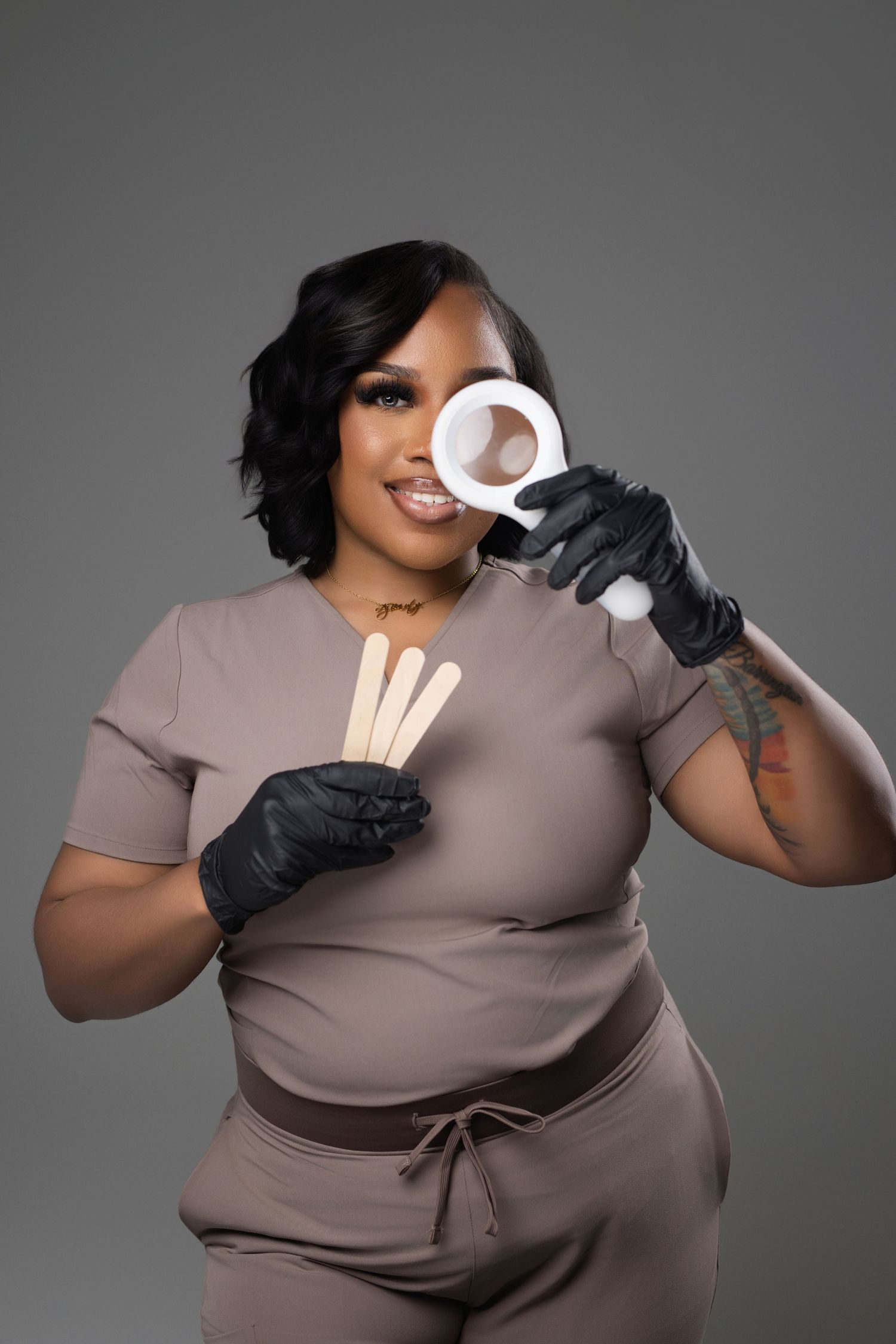 A plus-size Black woman in scrubs holds waxing supplies and smiles in a professional beauty studio.