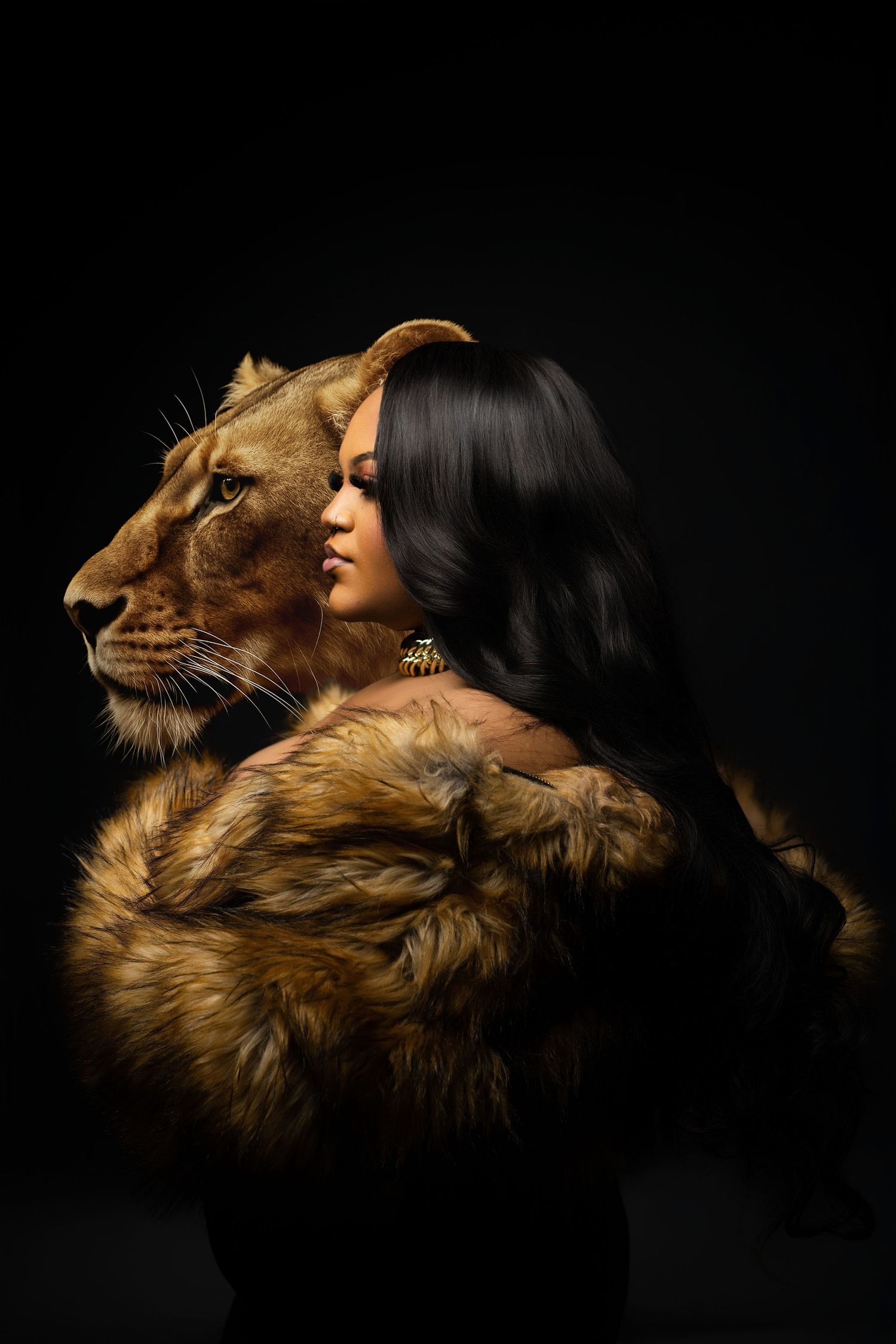 A woman in a fur coat poses dramatically with a lioness against a dark black background.
