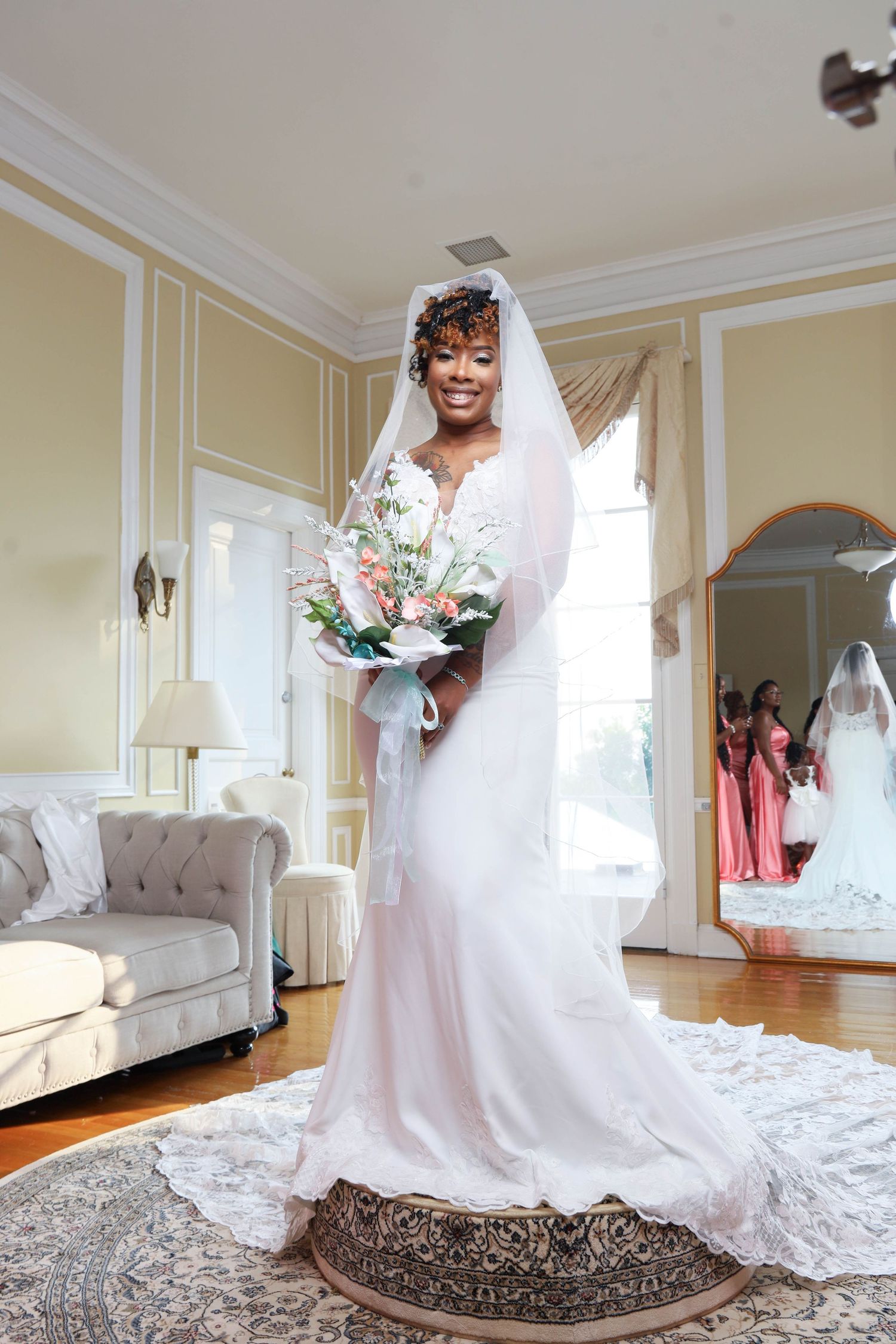 A smiling Black bride in a lace wedding gown and veil holds a floral bouquet in an elegant room.