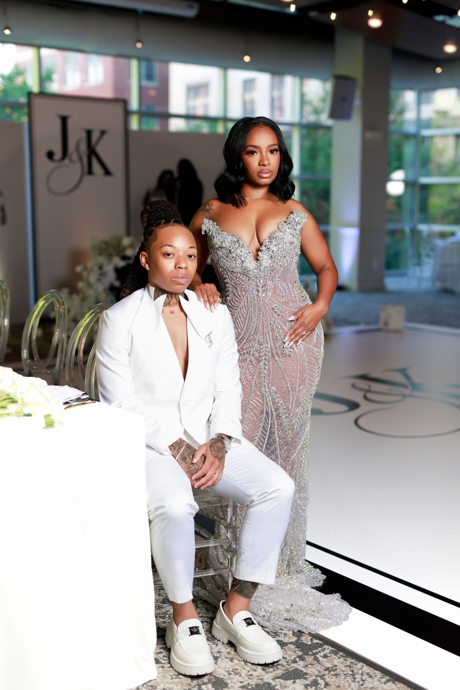 Same-sex couple poses at wedding reception, one in white suit, one in beaded gown.