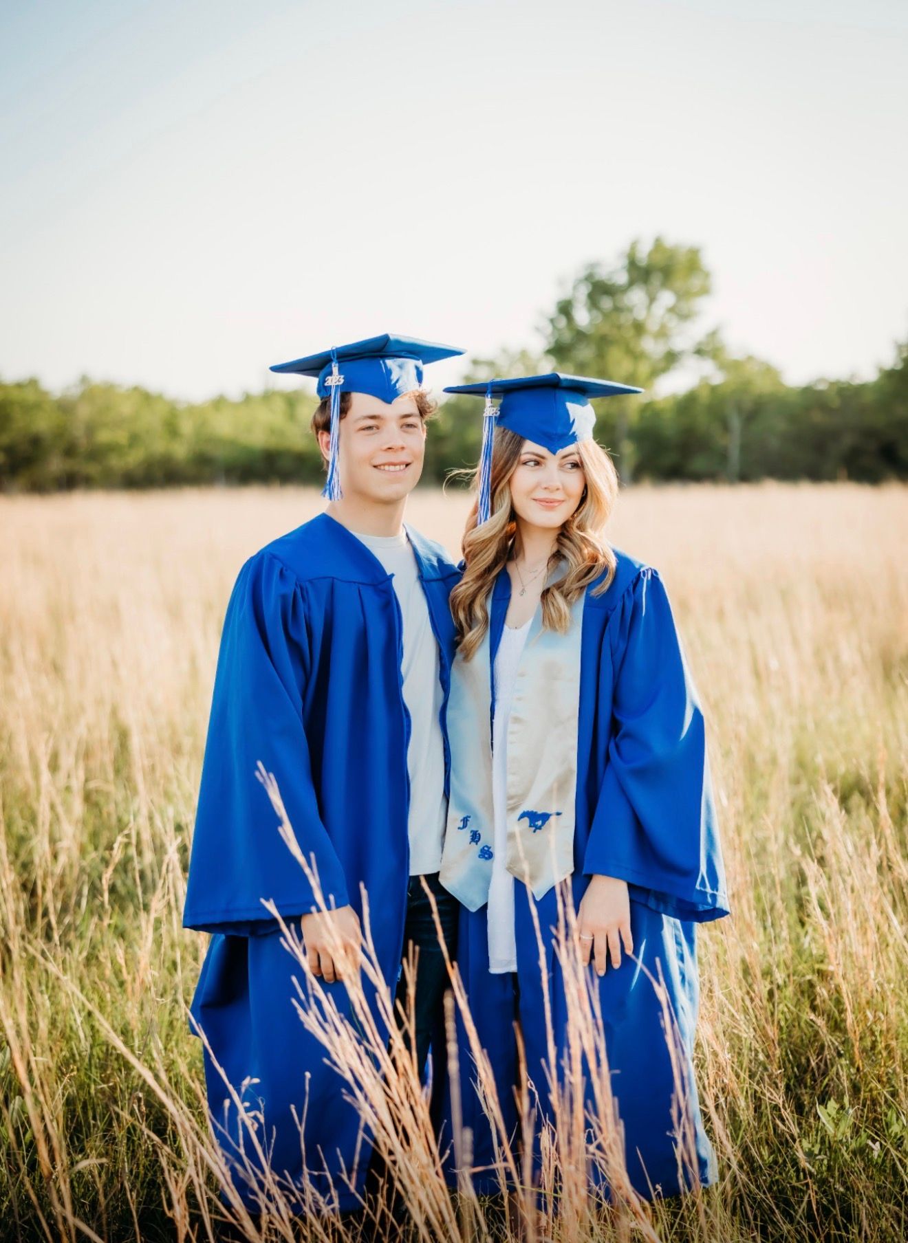 Two graduates in blue cap and gown posing together in a golden field during sunset.