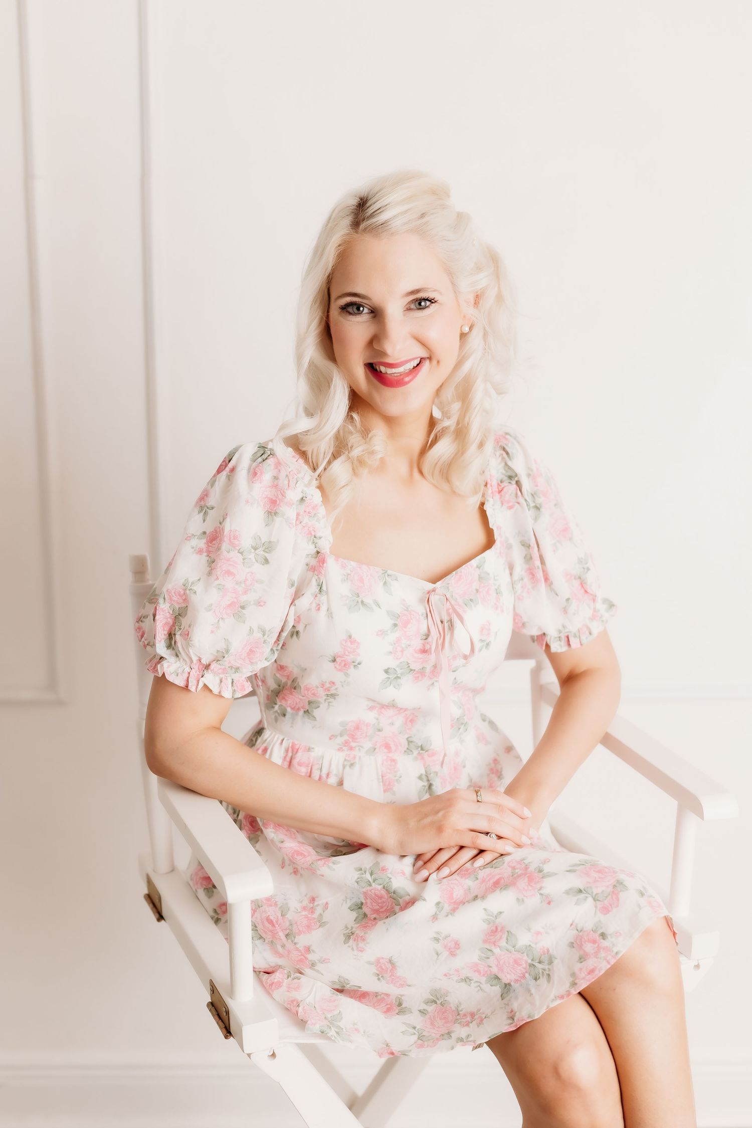 Smiling blonde woman in pink floral mini dress sitting on white chair in bright studio setting.