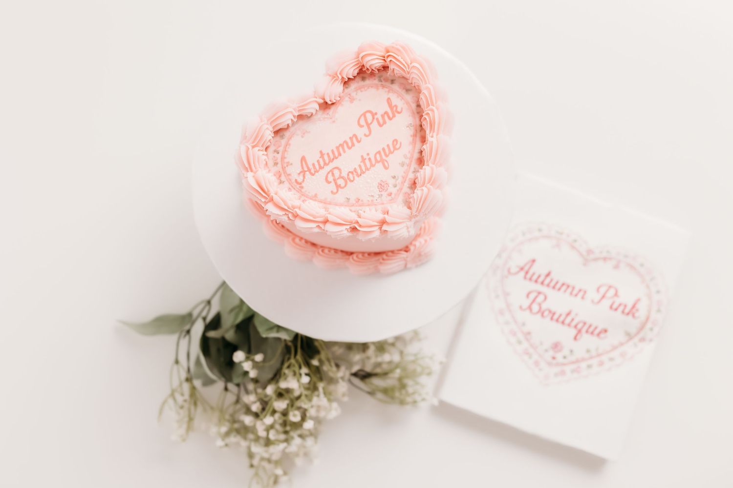 Pink heart-shaped Autumn Pink Boutique cake beside logo card and baby breath flowers on white surface.