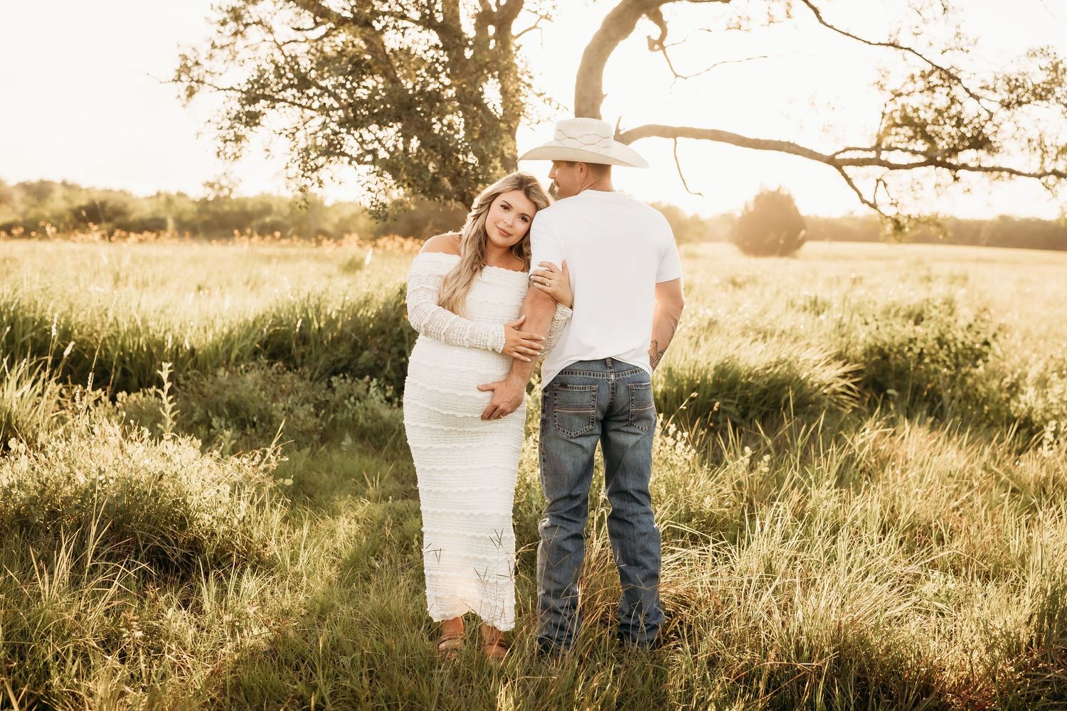 Expecting couple in golden hour field, woman in white lace dress and man in cowboy hat.