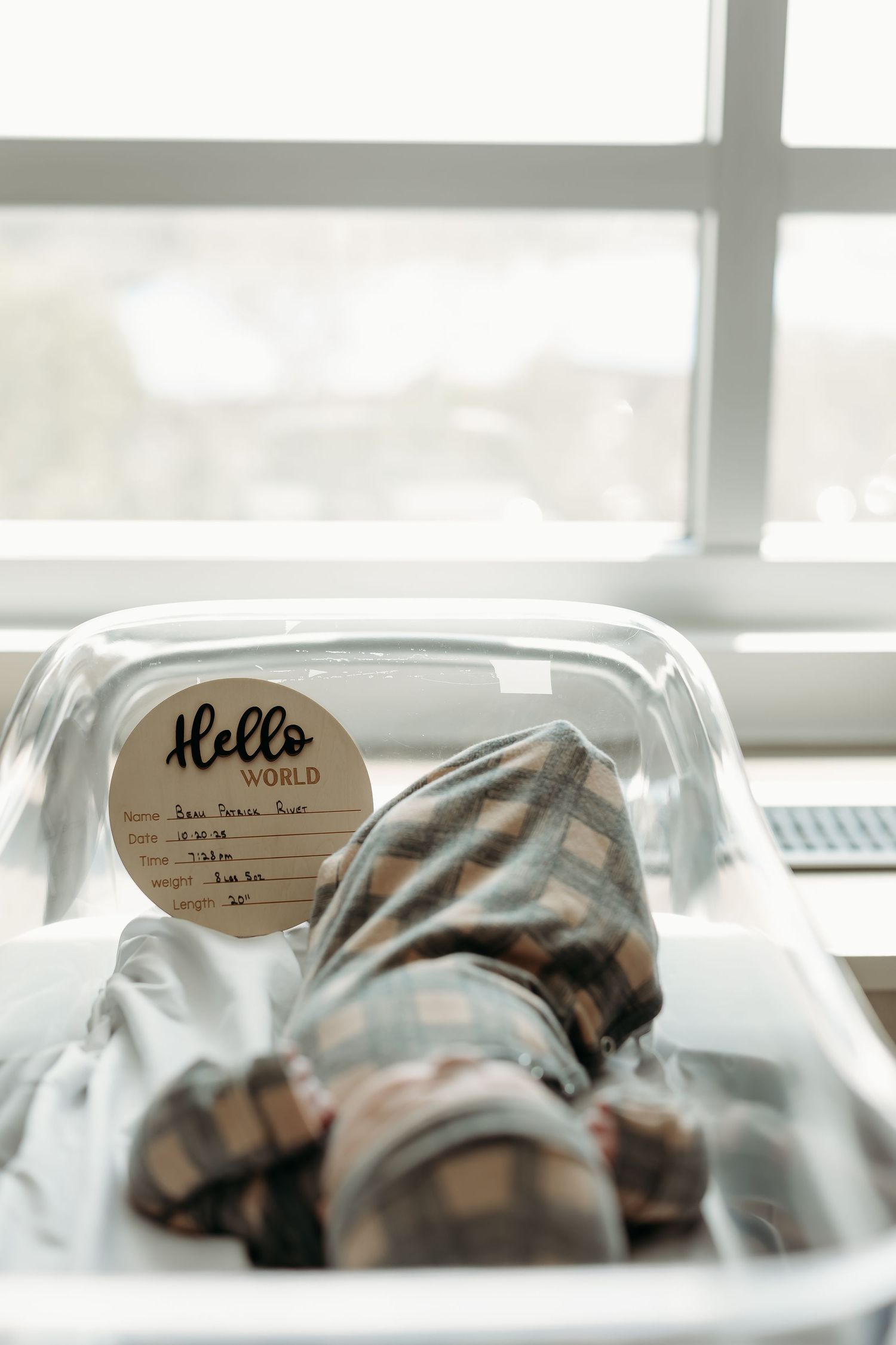 Newborn baby in plaid outfit lying in clear hospital bassinet with Hello World name tag.