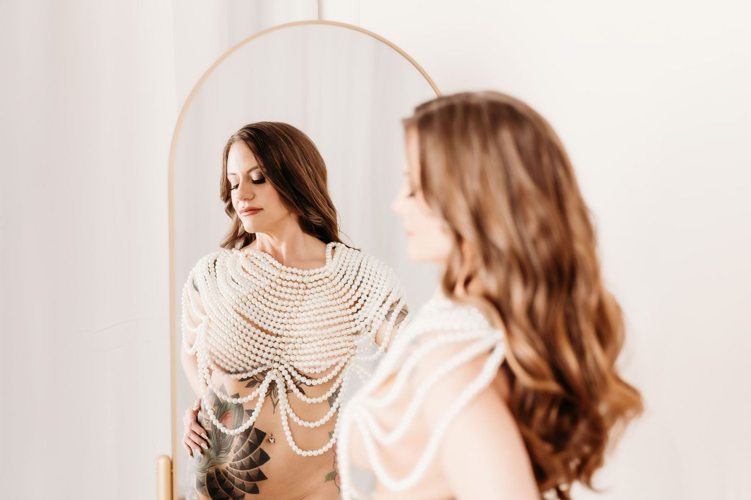 Woman in pearl body chain admires her reflection in a tall arched mirror.