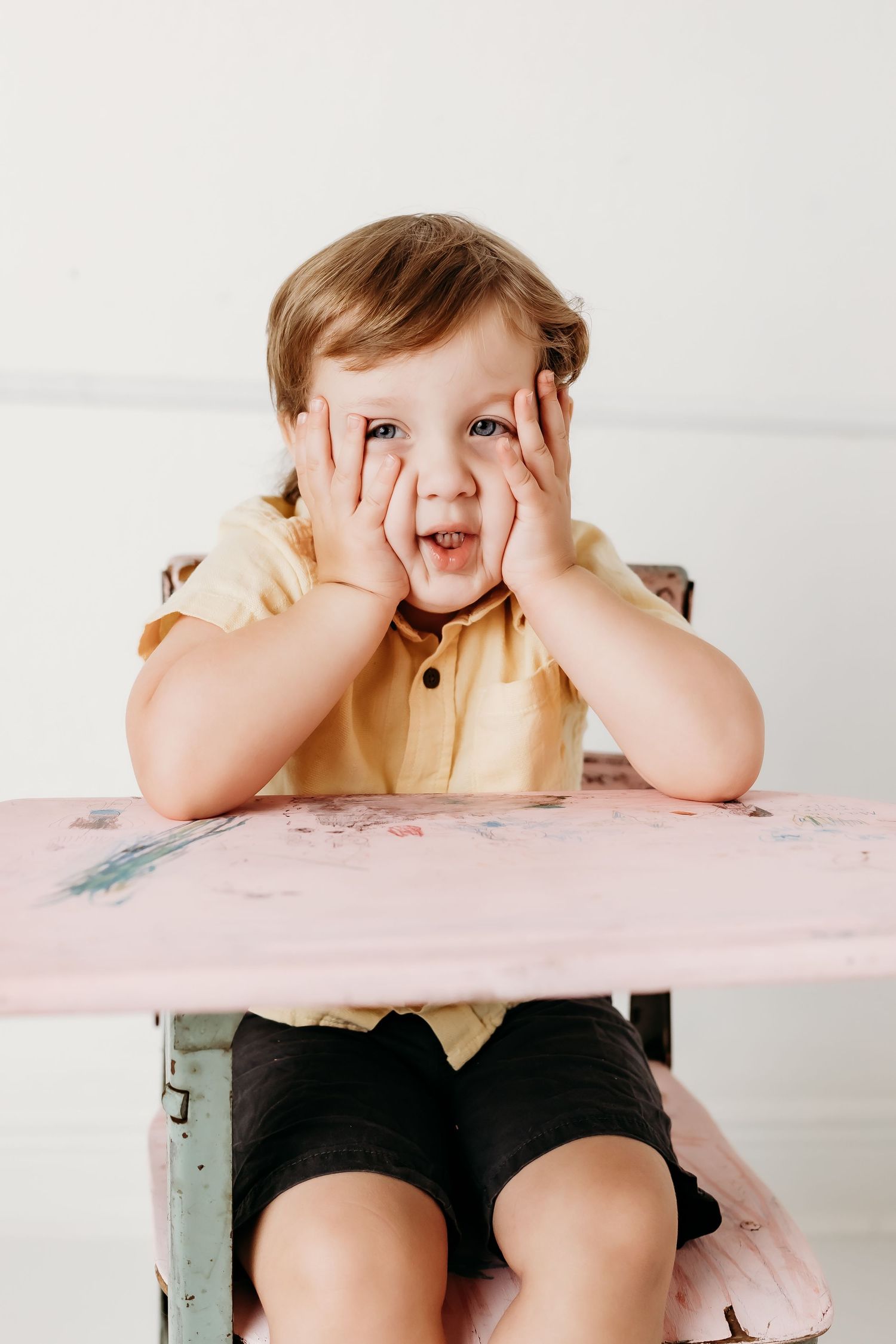 A young child in a yellow shirt makes a funny face with both hands on their cheeks at a vintage pink school desk.