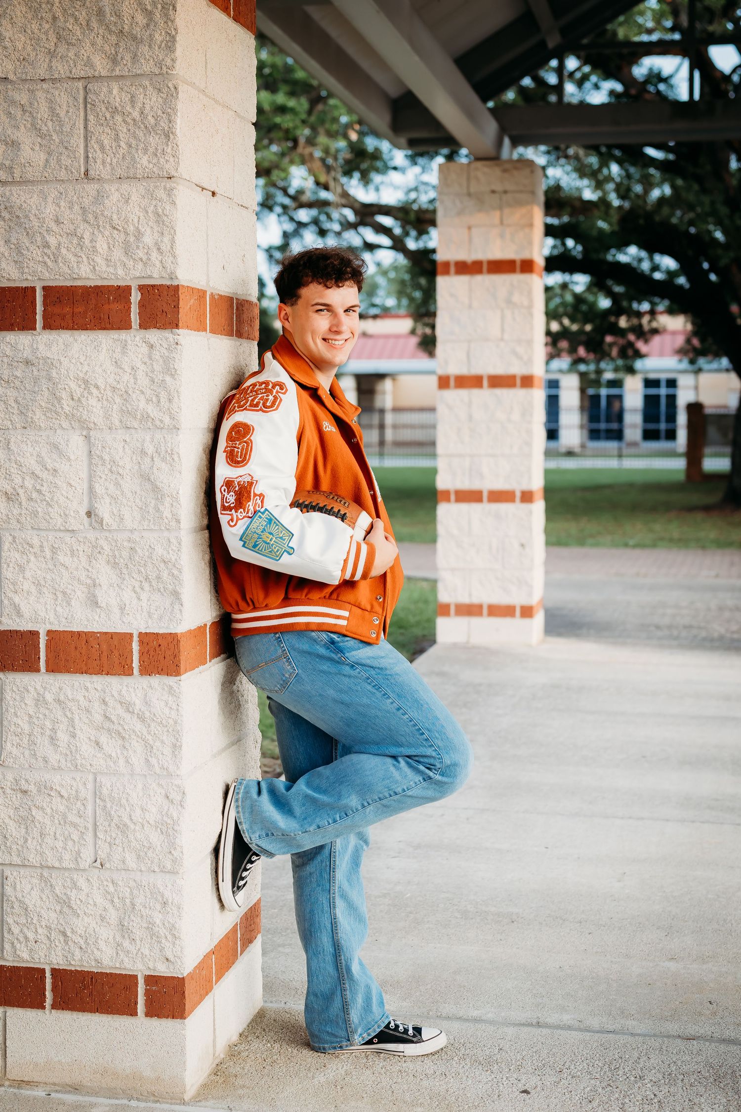 Smiling senior boy in orange letterman jacket leans against brick pillar outdoors.
