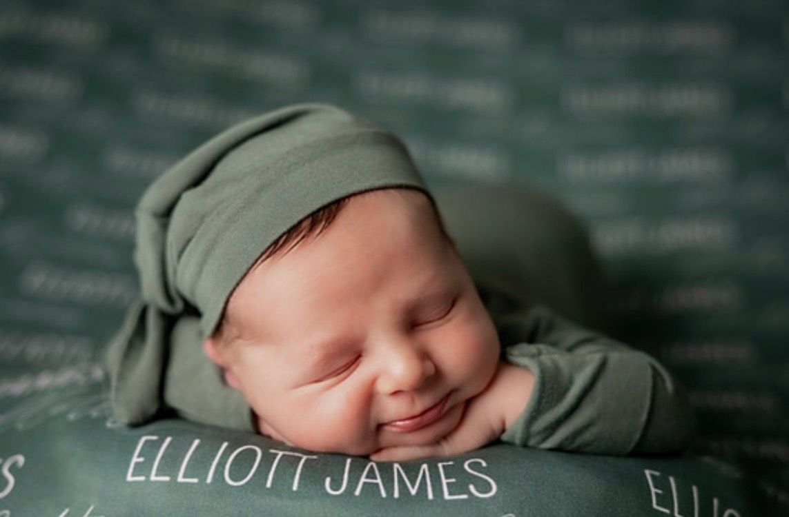 A smiling newborn baby dressed in green sleeps peacefully on a personalized blanket reading Elliott James.