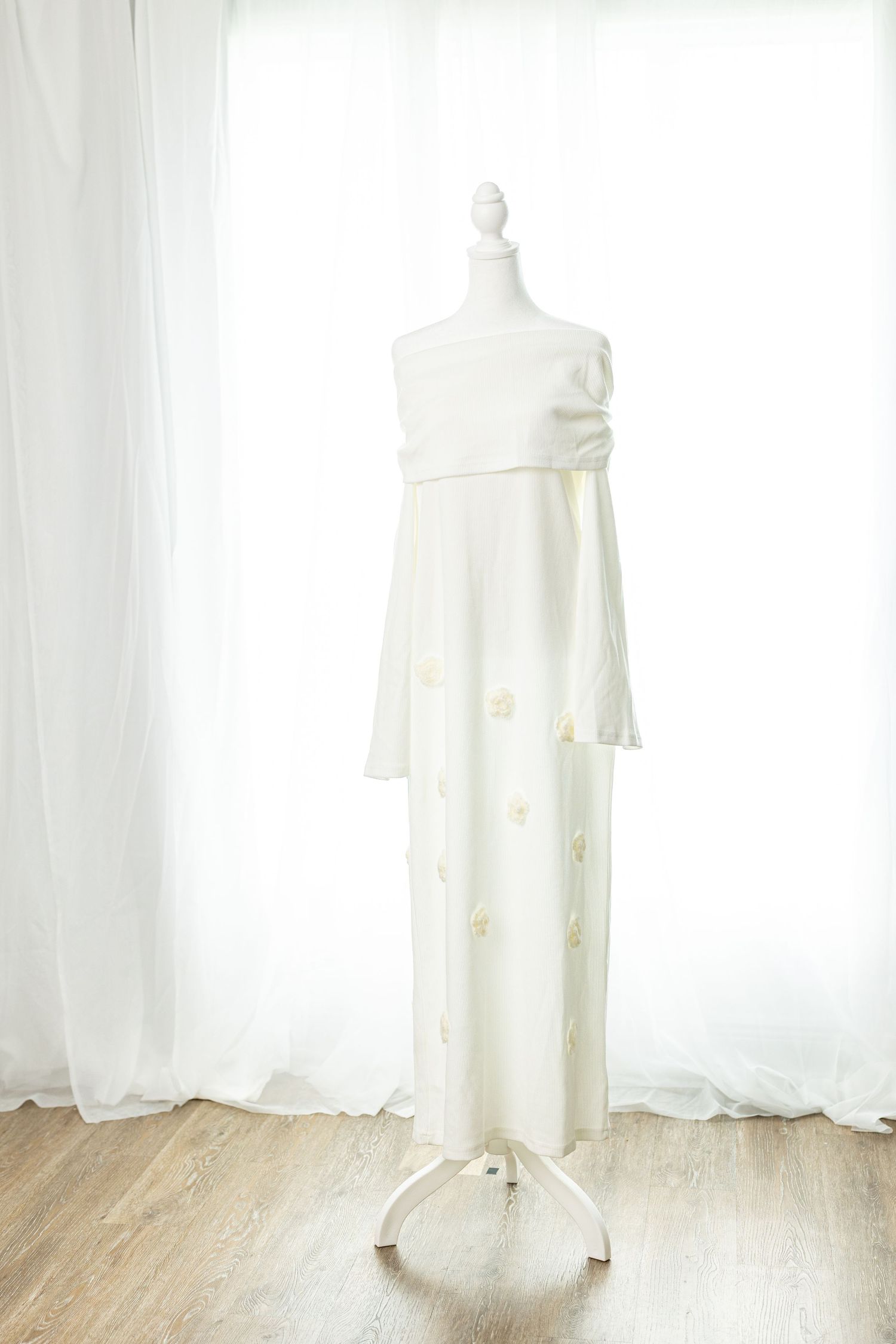 White off-shoulder maxi dress with gold floral embroidery displayed on a mannequin stand by sheer curtains.