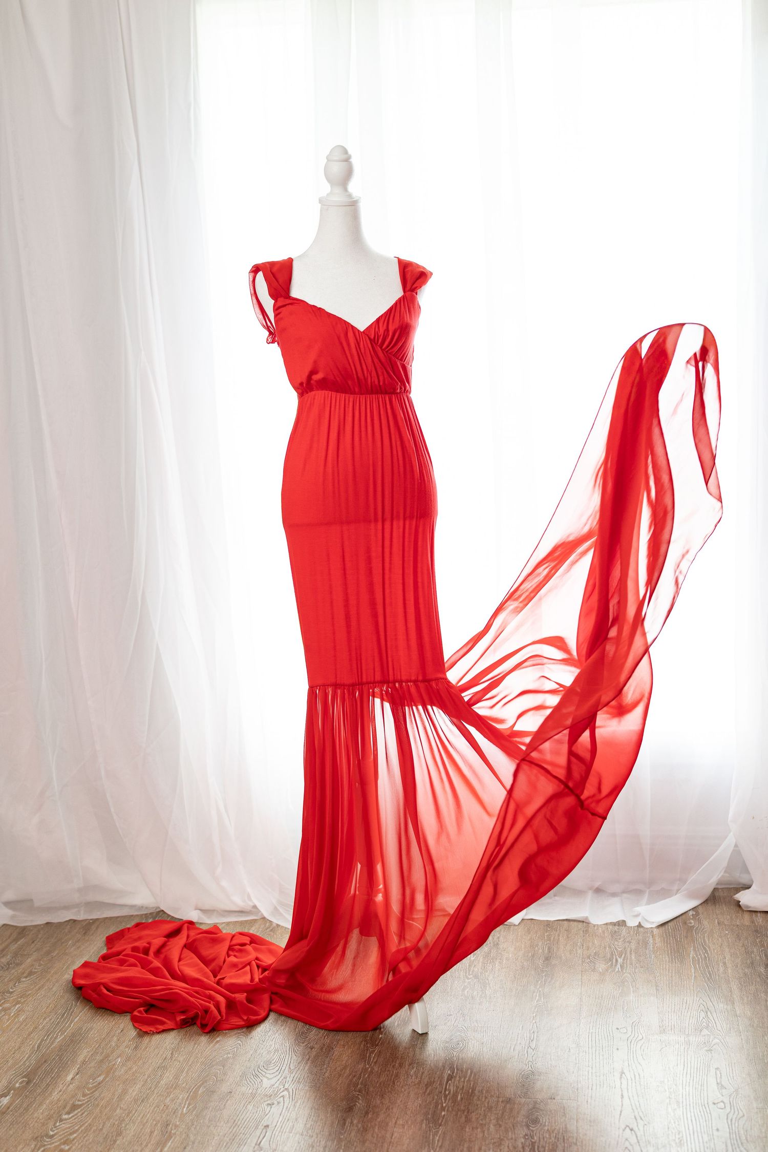 Elegant red chiffon maternity gown on a mannequin with flowing train, displayed against white curtains.