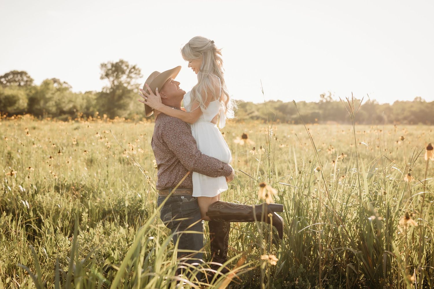 Man lifting woman in white dress in a glowing sunlit wildflower meadow during golden hour.