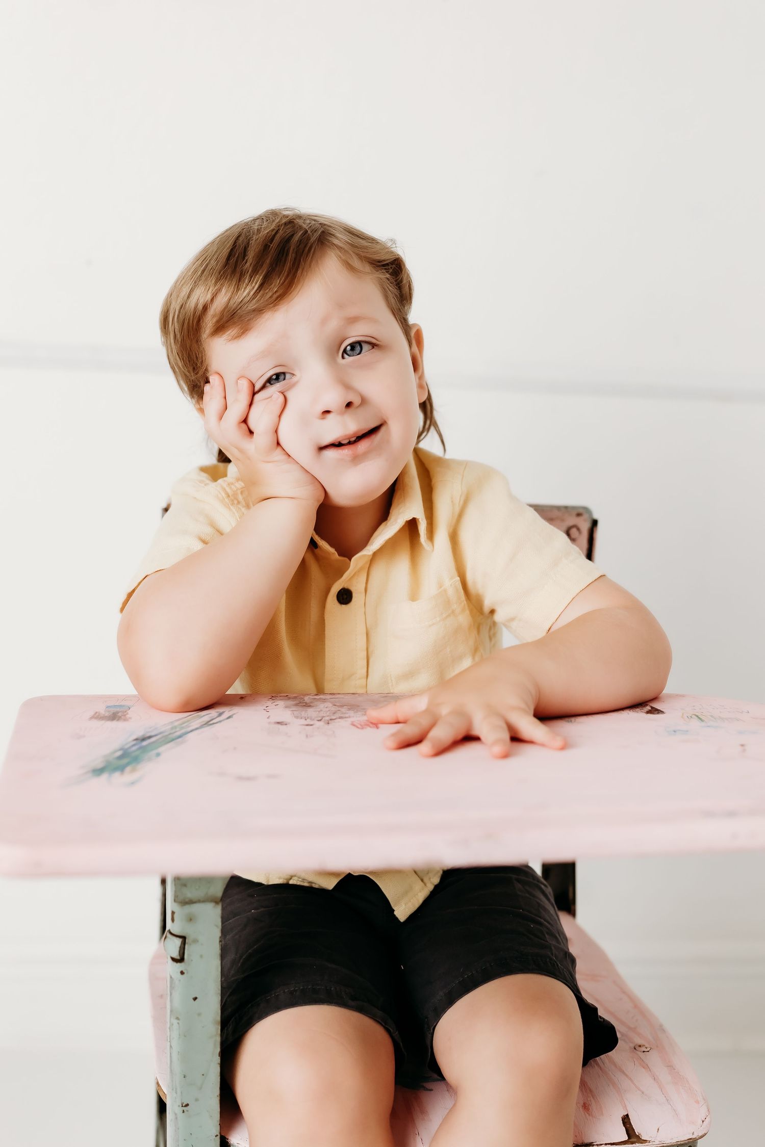 A young child in a yellow shirt smiles while resting their cheek on their hand at a vintage pink school desk.