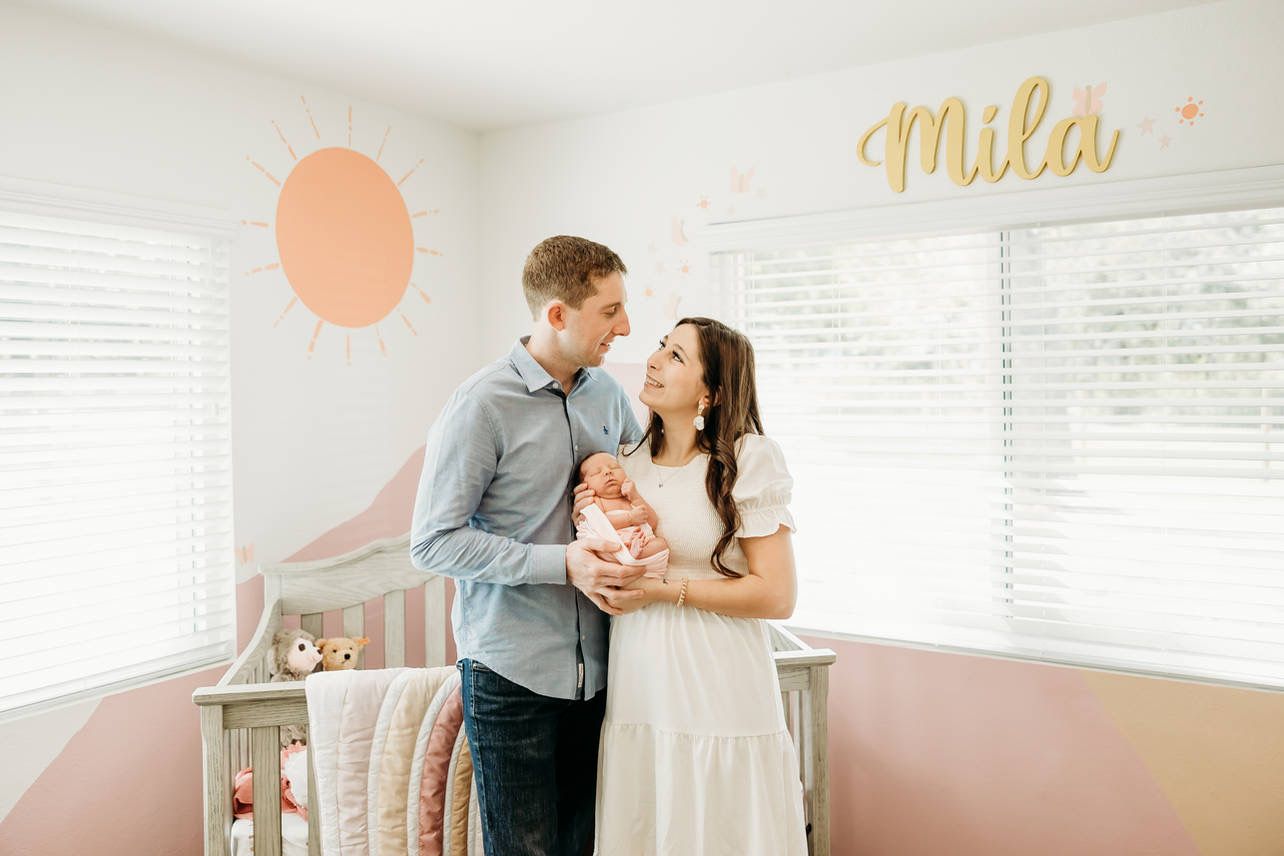 Parents smile at newborn baby in pink nursery with Mila name sign and sun wall mural.