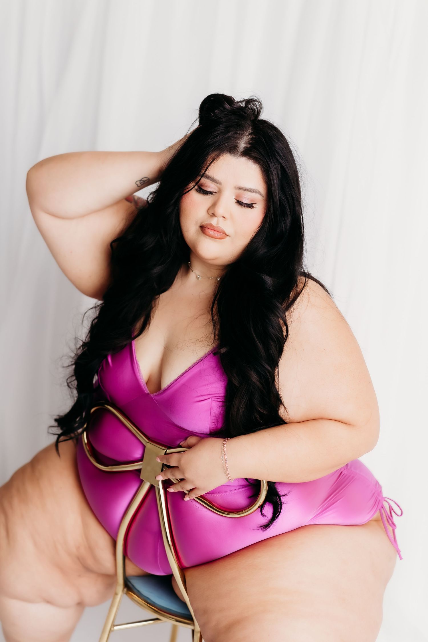 Plus size woman in magenta satin swimsuit posing on gold chair against white backdrop.