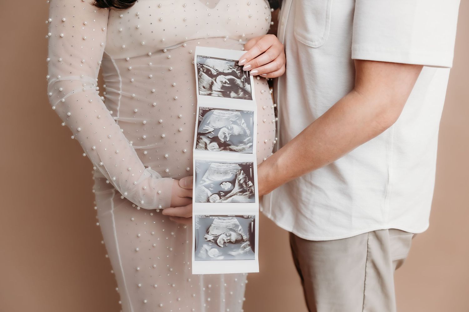 Pregnant woman in pearl dress and partner holding ultrasound photos against beige background.