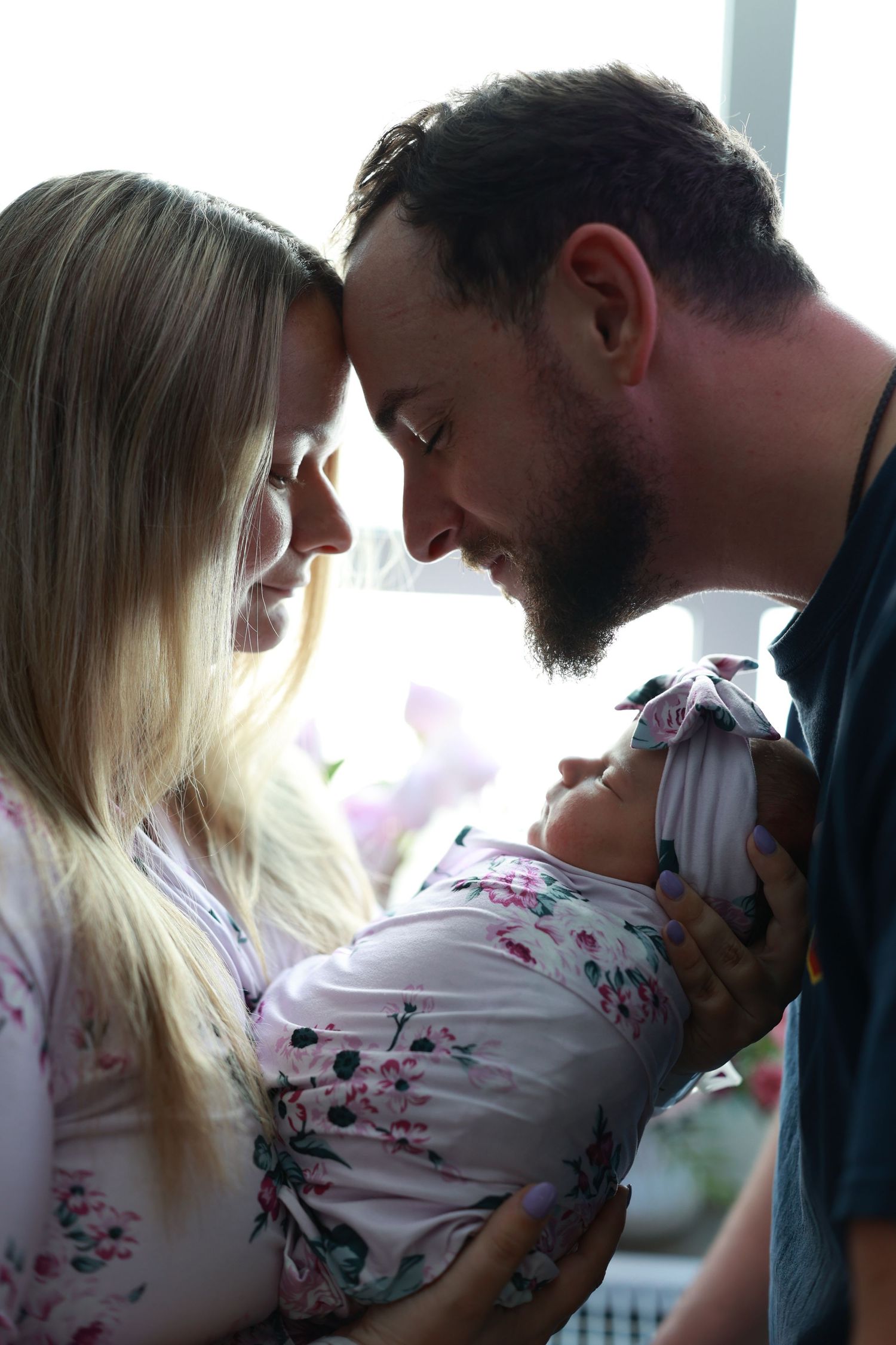 New parents lovingly touch foreheads together while holding their swaddled newborn baby near window.