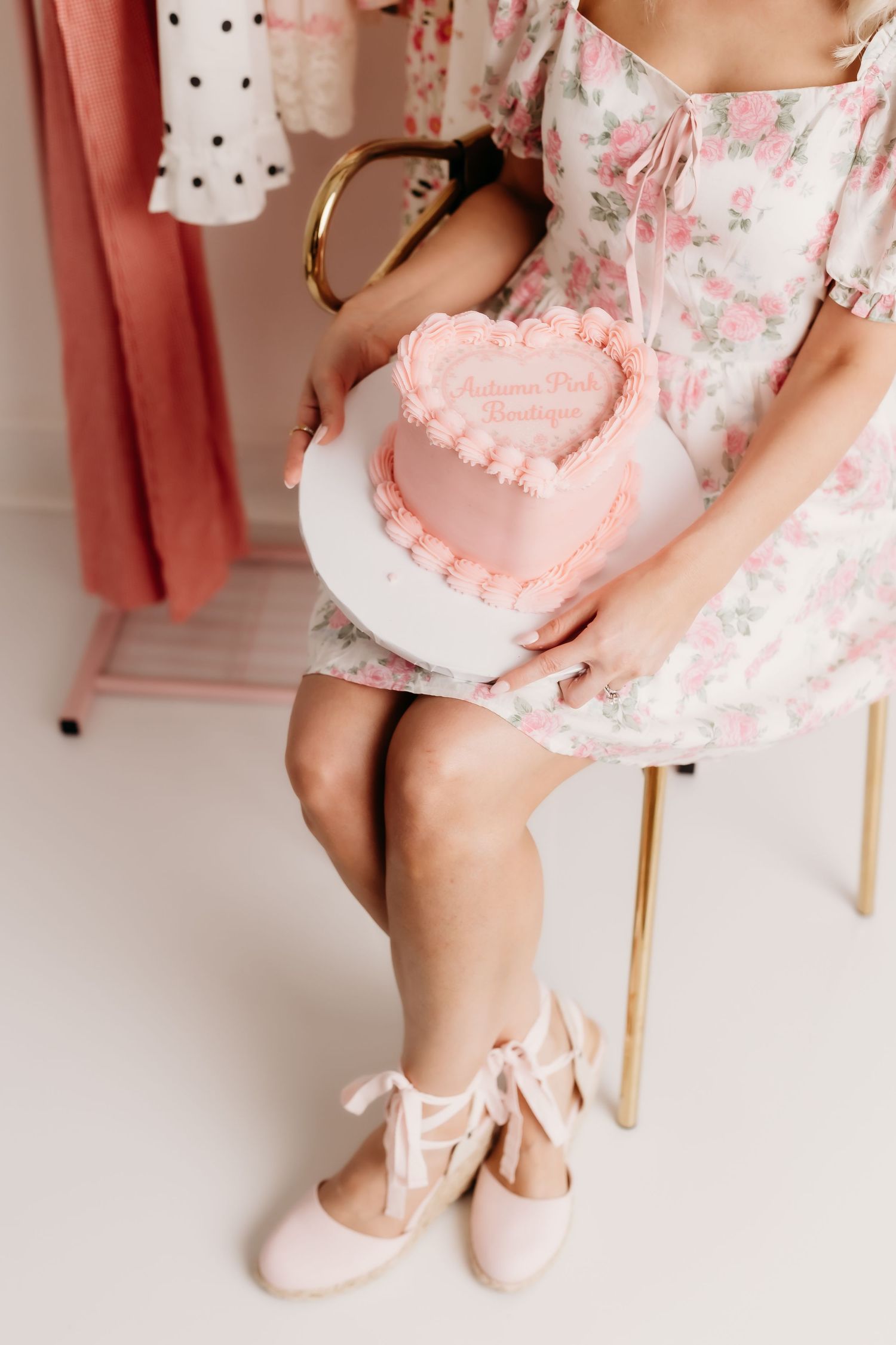 Woman in floral dress holding pink heart-shaped cake while seated in gold chair with ballet flats.