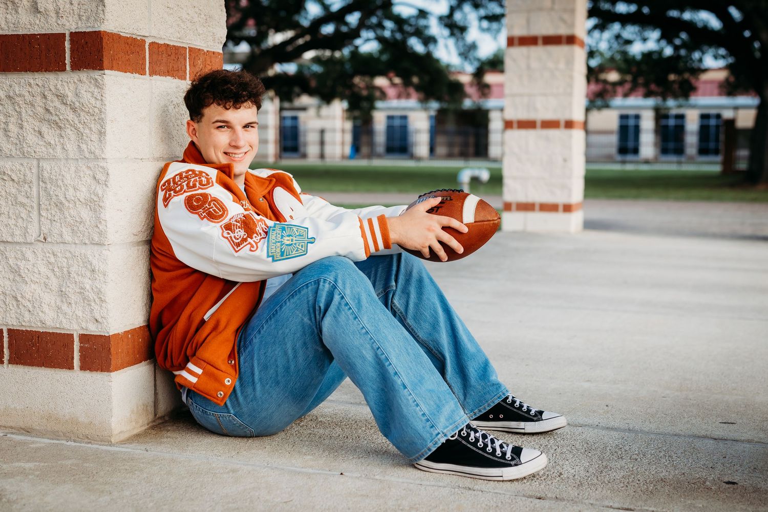 Senior boy in orange letterman jacket sits against brick column holding football outdoors.