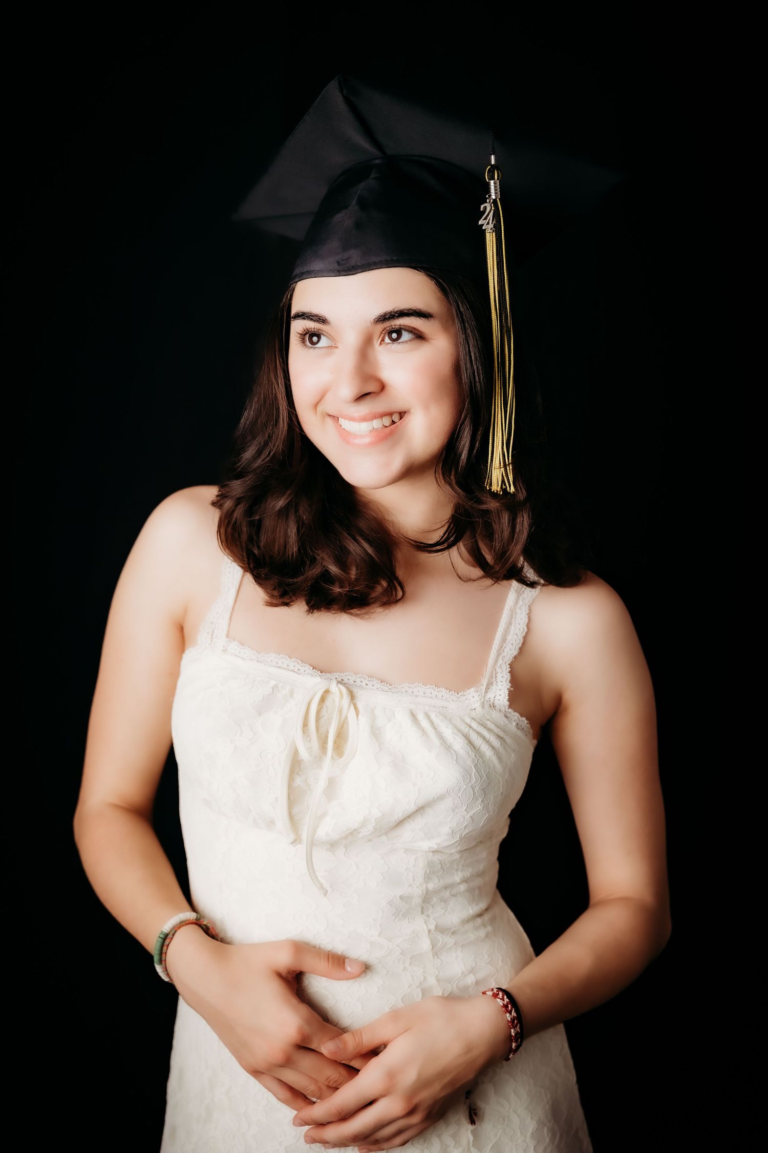 Smiling brunette in graduation cap and white dress posing against a dark black background.