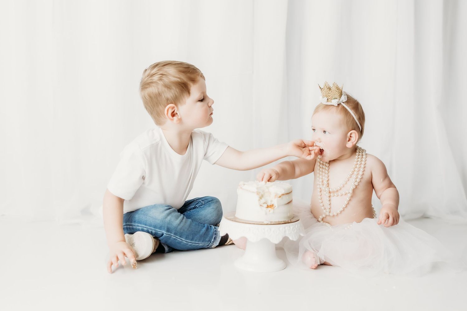 A young boy feeds cake to a baby girl wearing a pearl necklace and crown during a first birthday cake smash photo session.