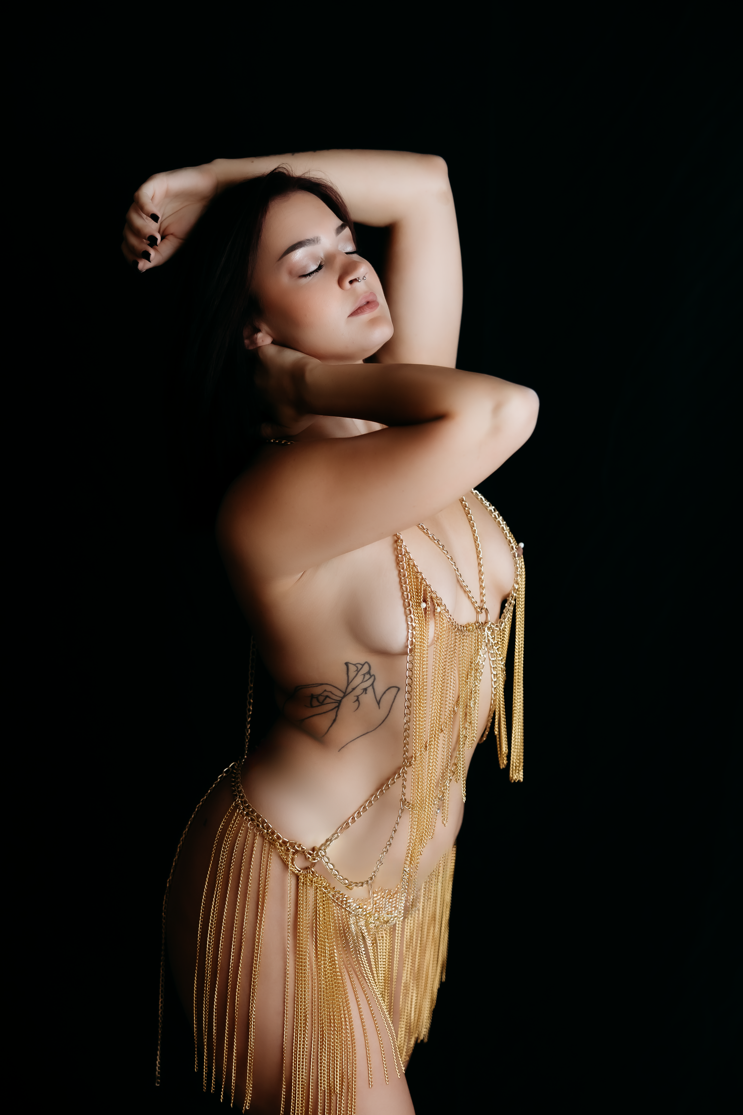 Woman posing in gold fringe body jewelry with tattoo on back against black background.