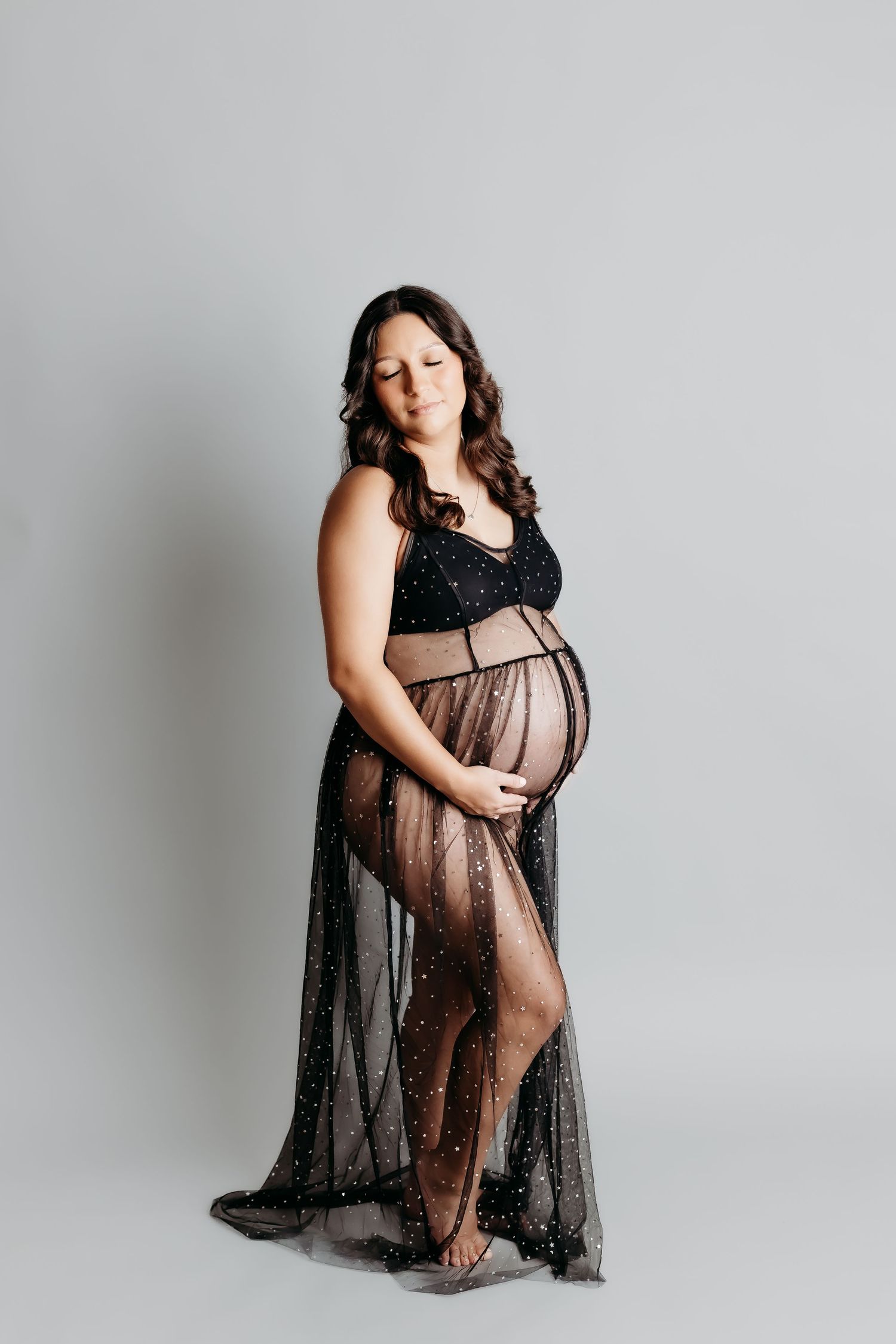 Pregnant woman in black sequined bodysuit and sheer skirt cradling baby bump during maternity photoshoot.