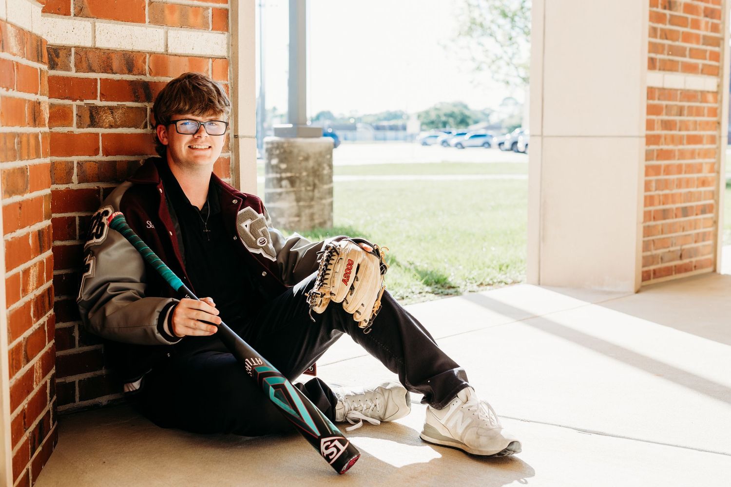 Teen boy with glasses sits against brick wall holding baseball bat and glove for senior photos.