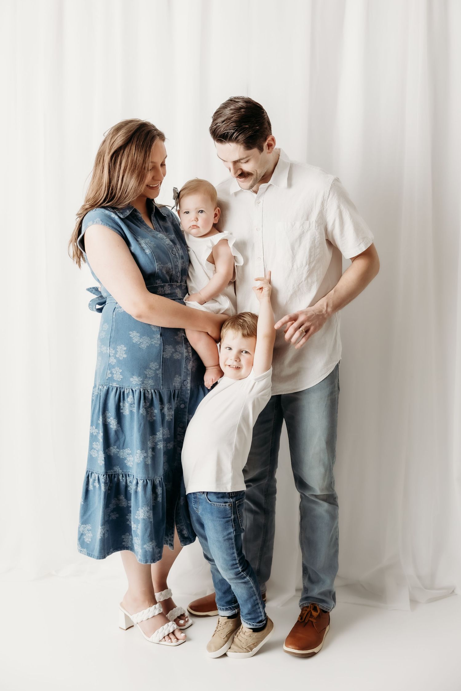 A happy family of four poses in a bright studio, with parents holding a baby and a young child reaching up.